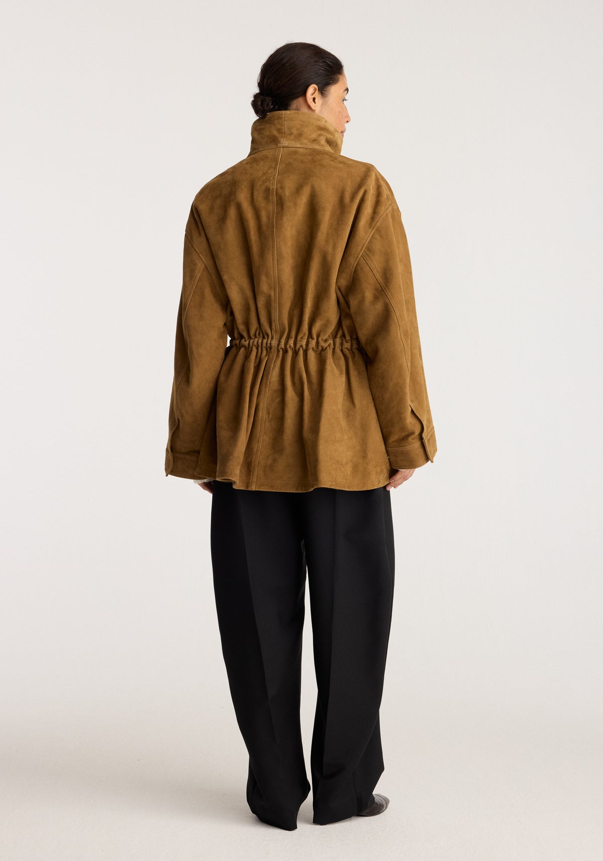 Sculptural suede jacket | sage shown from the back with a gathered waist high collar long sleeves and relaxed sculptural fit styled with black trousers