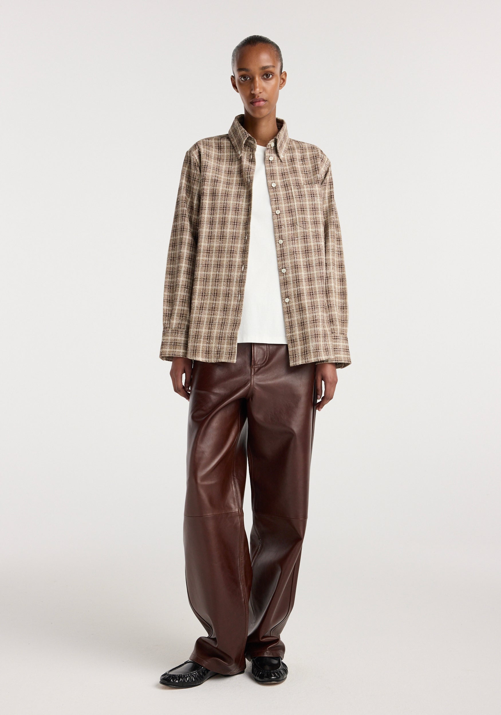 Woman wearing an Oxford bouclé check shirt | boucle check in beige and brown tones, paired with white top and dark brown leather pants, standing against a plain background.