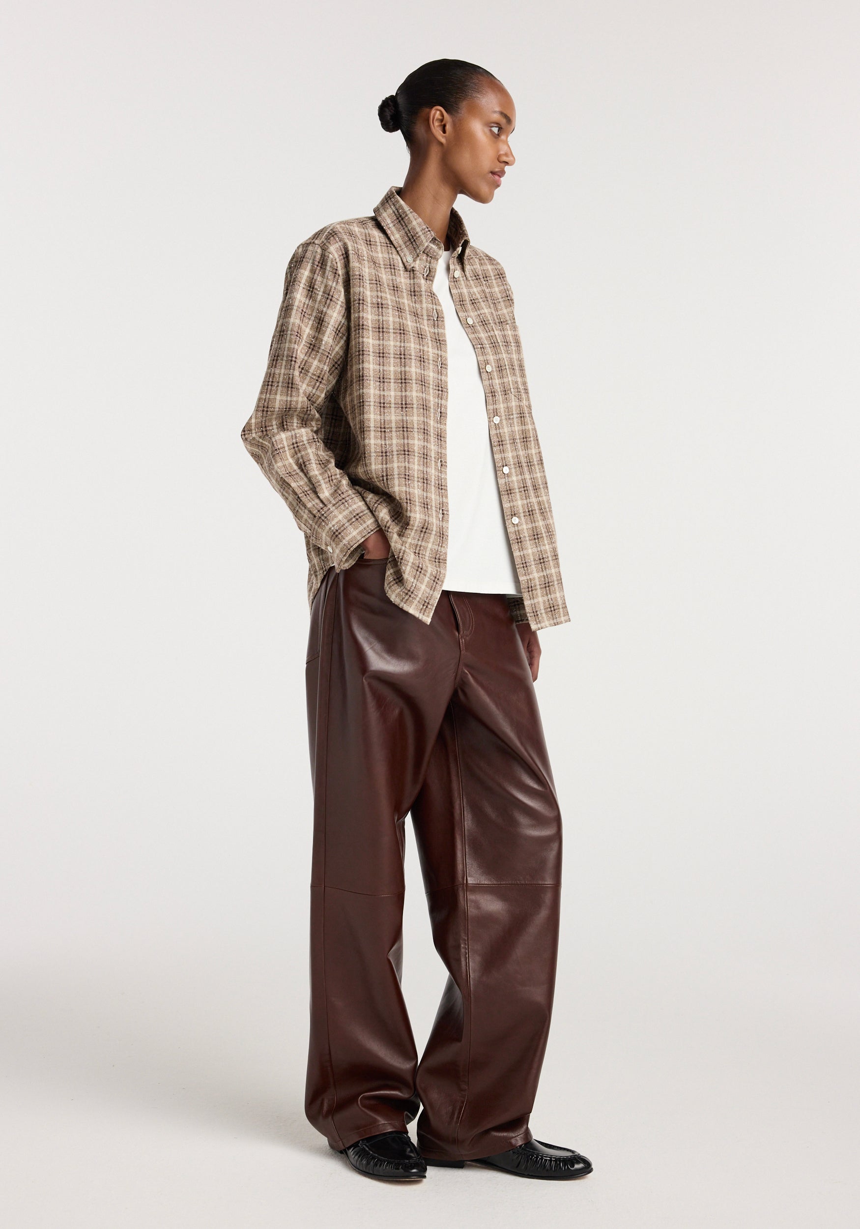 Woman wearing an Oxford bouclé check shirt | boucle check in neutral tones, paired with a white top and wide-leg brown leather pants, standing against a plain background.