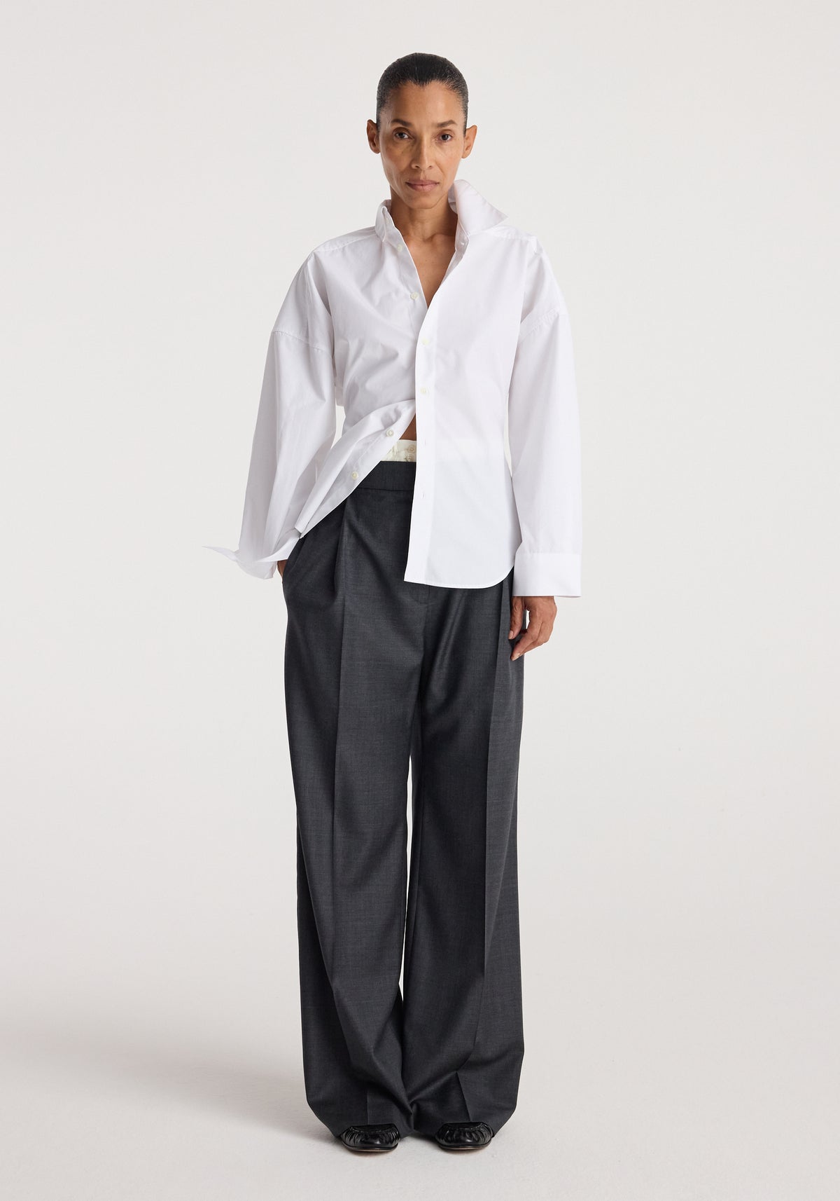 Model wearing a long-sleeve Poplin uneven collar shirt | optic white with a partially open front, paired with loose dark gray pants against a plain background.