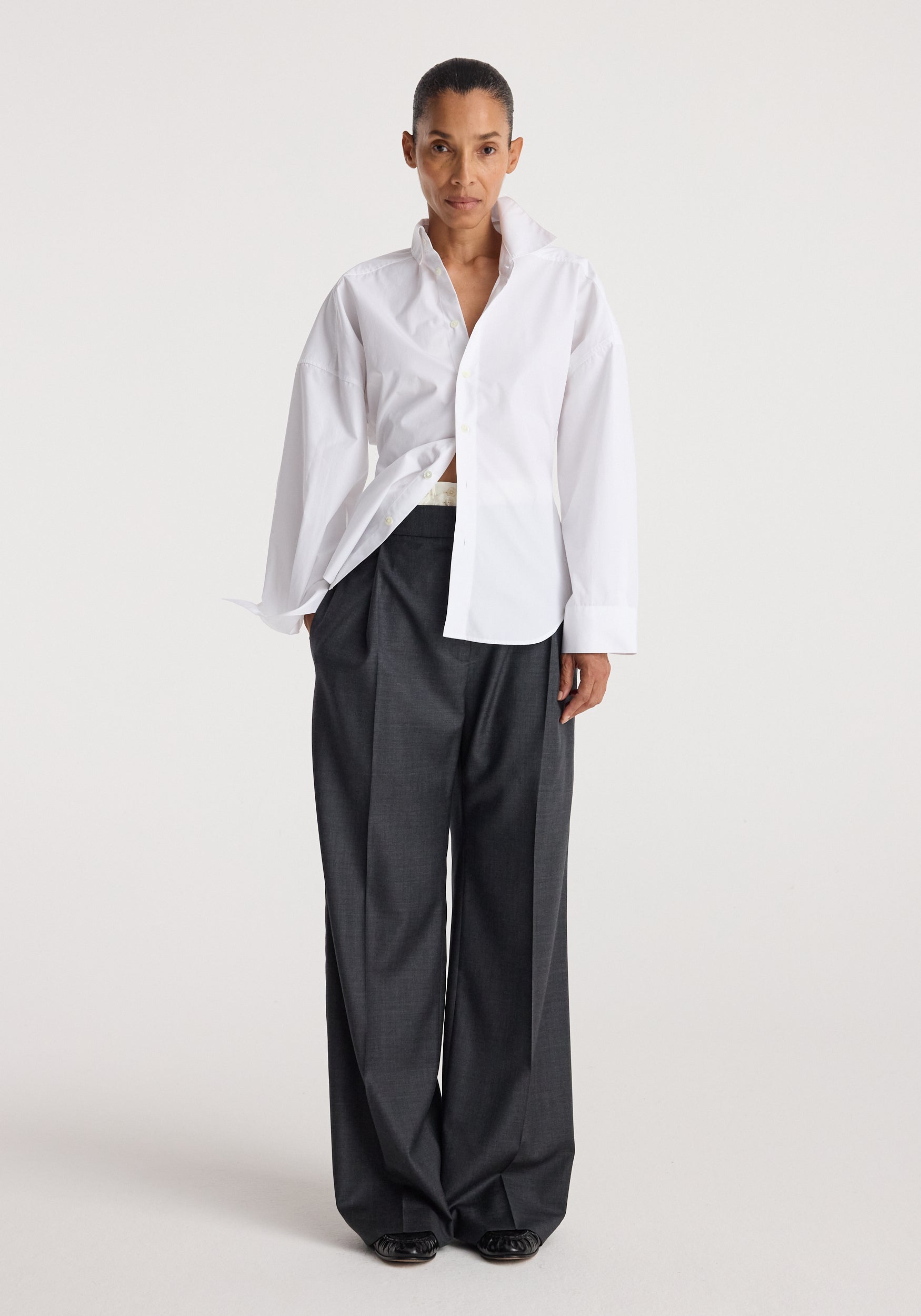 Model wearing a long-sleeve Poplin uneven collar shirt | optic white with a partially open front, paired with loose dark gray pants against a plain background.
