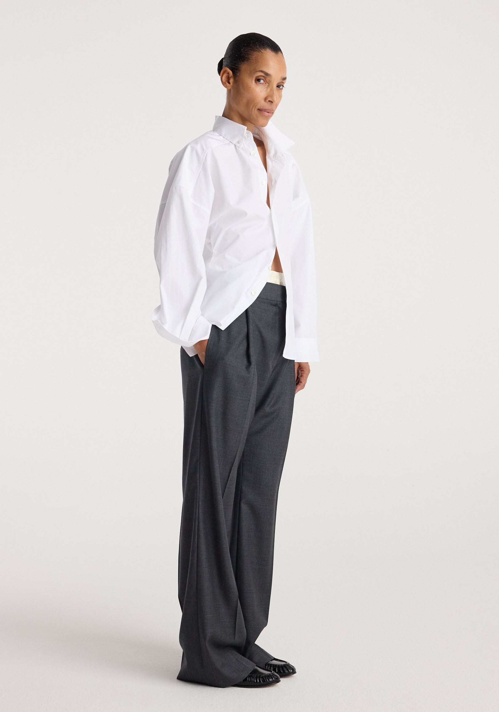 Woman wearing a loose-fitting Poplin uneven collar shirt | optic white paired with wide-leg dark gray trousers and black shoes against a plain background.