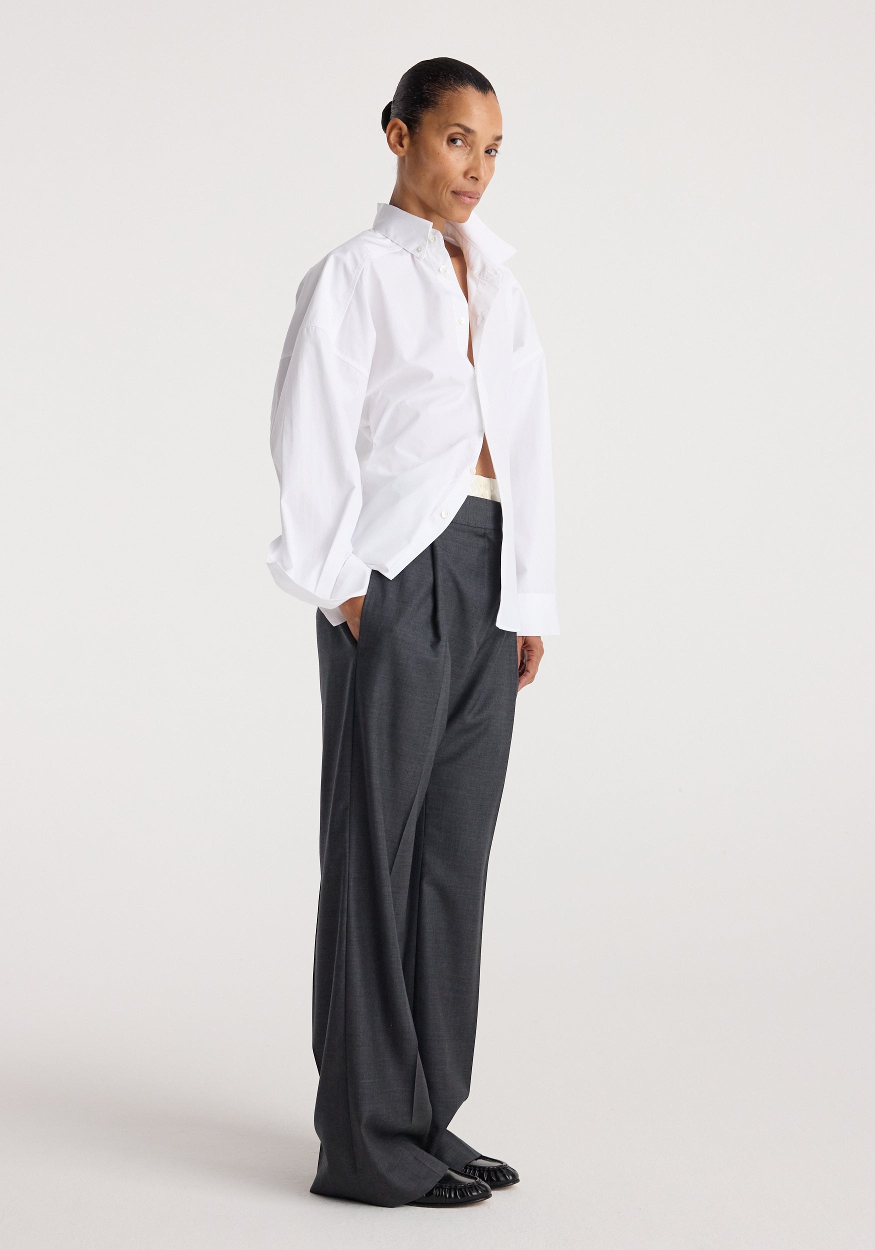 Woman wearing a loose-fitting Poplin uneven collar shirt | optic white paired with wide-leg dark gray trousers and black shoes against a plain background.