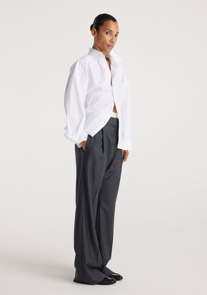Woman wearing a loose-fitting Poplin uneven collar shirt | optic white paired with wide-leg dark gray trousers and black shoes against a plain background.