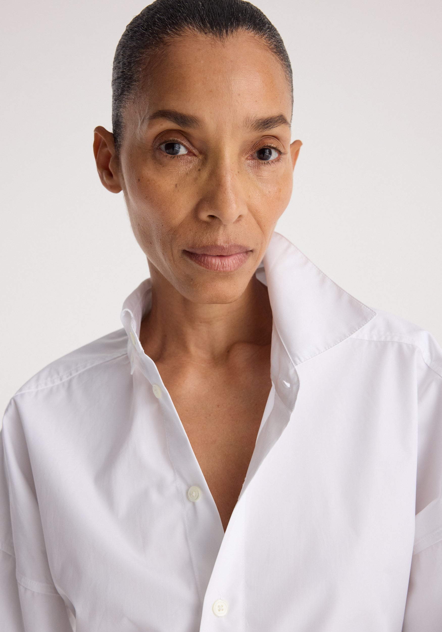 Close-up of a woman wearing a crisp white button-up Poplin uneven collar shirt | optic white with the collar turned up against a plain light background.