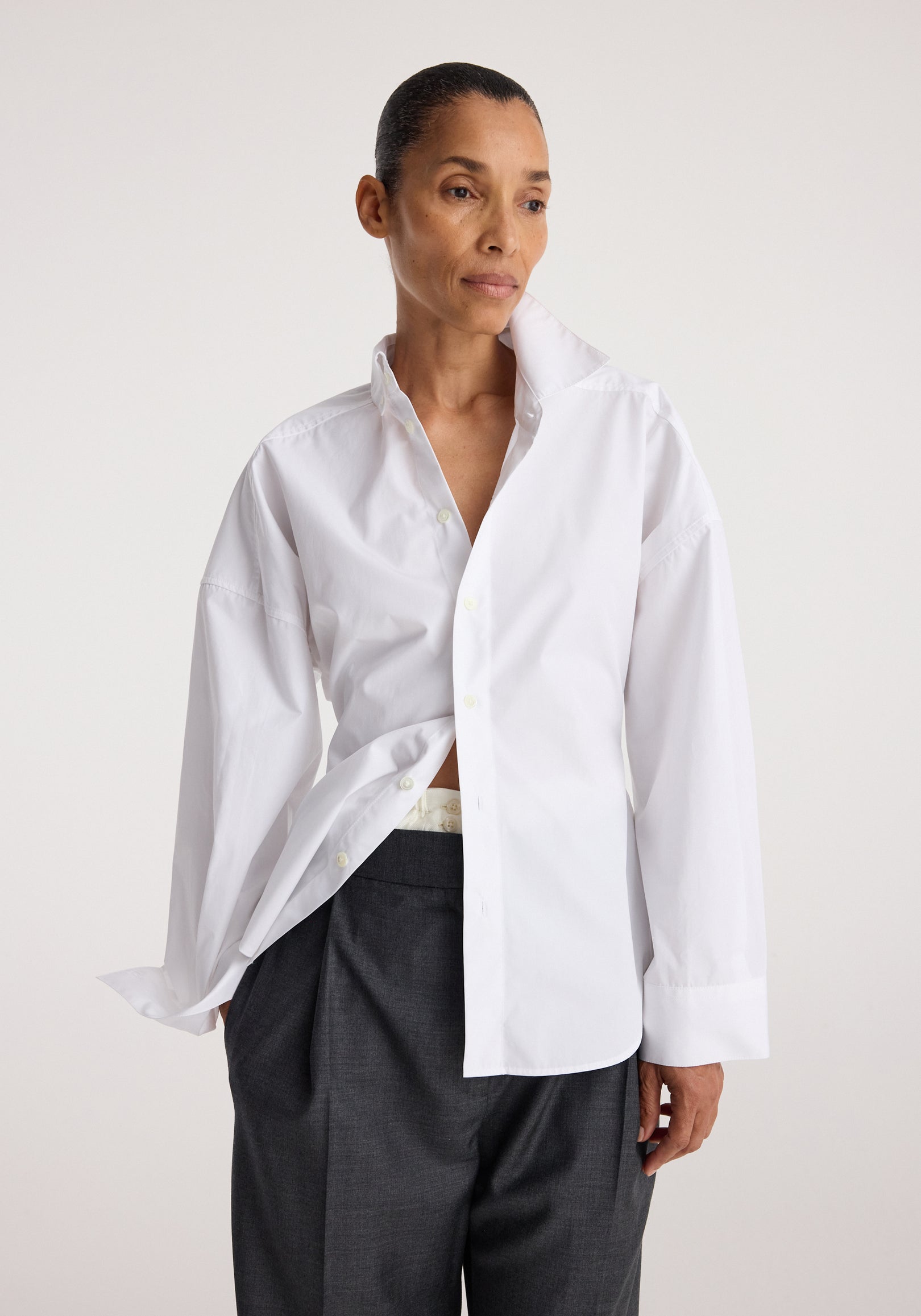 Woman wearing a loose-fitting Poplin uneven collar shirt | optic white with long sleeves and dark trousers, posing against a plain light background.