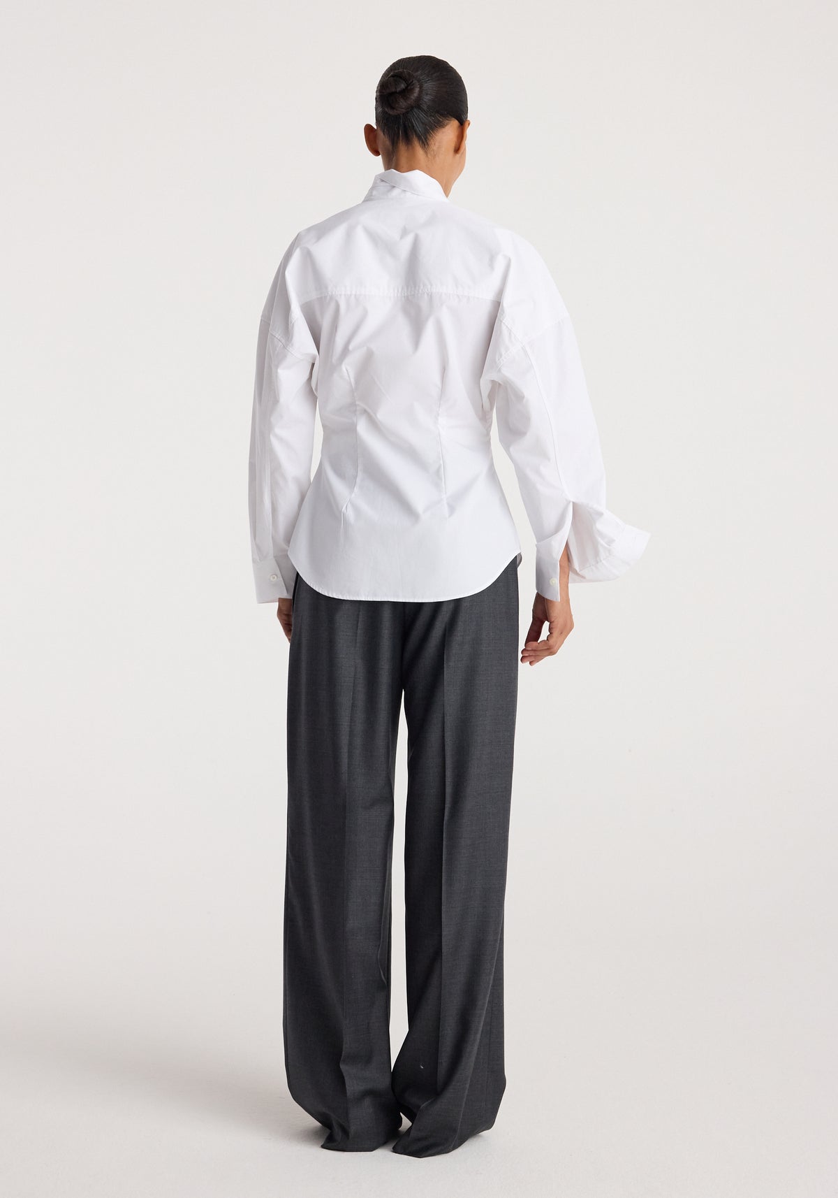 Model wearing a Poplin uneven collar shirt | optic white, shown from the back with long sleeves and a curved hem, paired with dark wide-leg trousers.