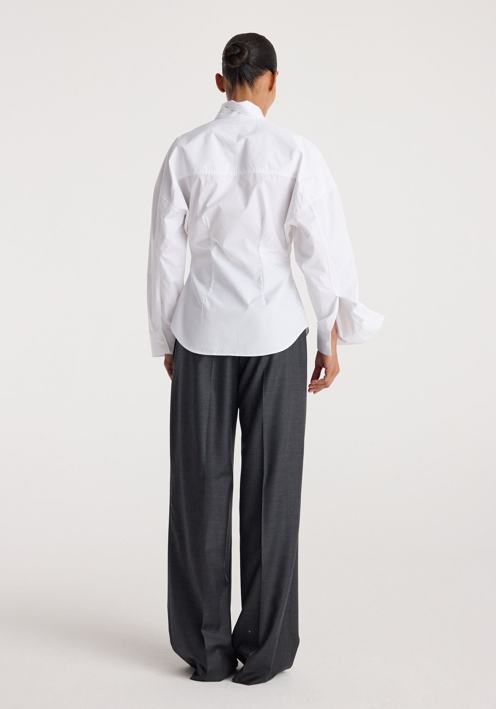 Model wearing a Poplin uneven collar shirt | optic white, shown from the back with long sleeves and a curved hem, paired with dark wide-leg trousers.