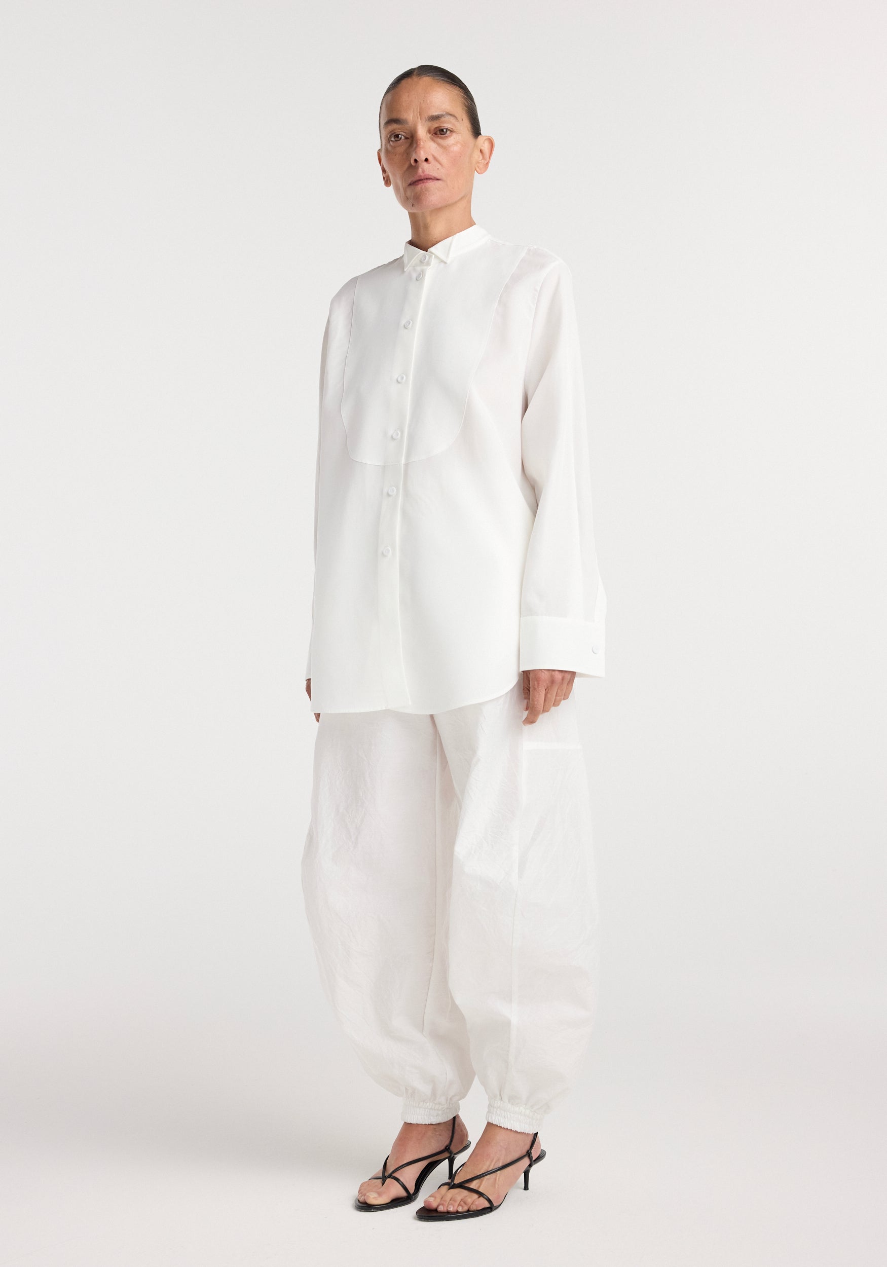 A model wearing a long-sleeve tuxedo shirt | optic white with a stand-up collar and concealed buttons, paired with loose white pants and black strappy heels.