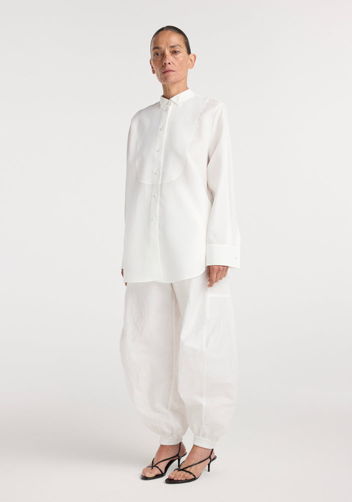 A model wearing a long-sleeve tuxedo shirt | optic white with a stand-up collar and concealed buttons, paired with loose white pants and black strappy heels.
