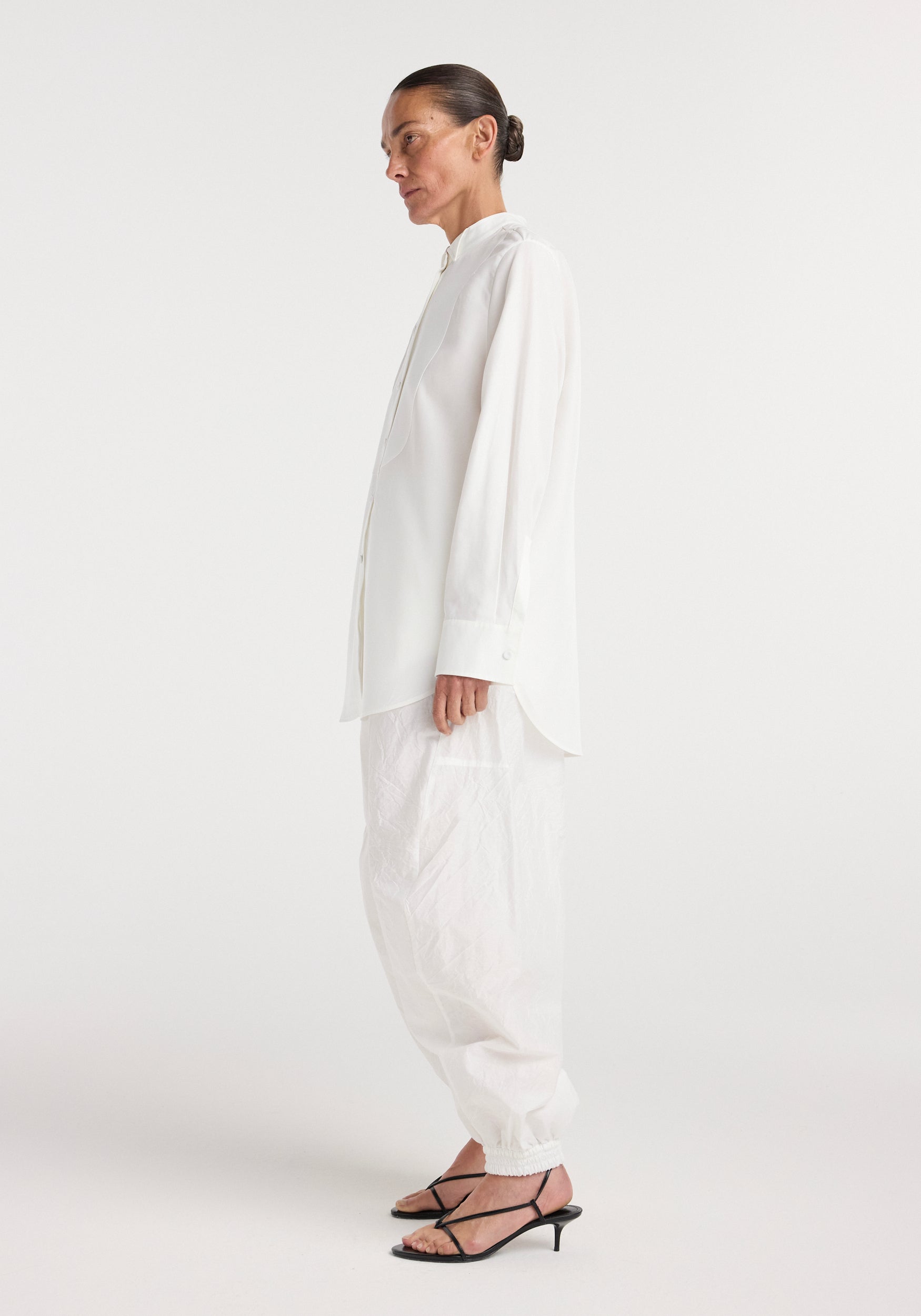 Side view of a person wearing a loose-fitting tuxedo shirt | optic white with long sleeves and buttoned cuffs, paired with casual white pants and black strappy low-heeled sandals.