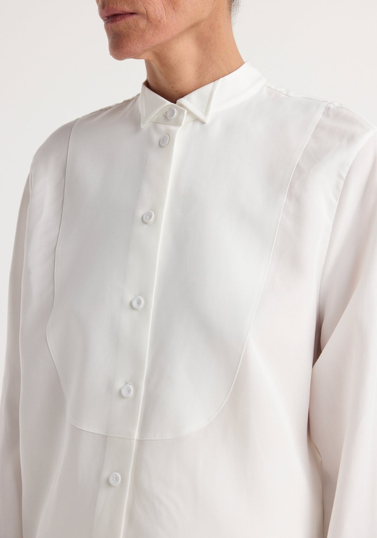 Close-up of a person wearing a classic tuxedo shirt | optic white featuring a pleated bib front and buttoned collar, highlighting the smooth fabric and fine detailing.