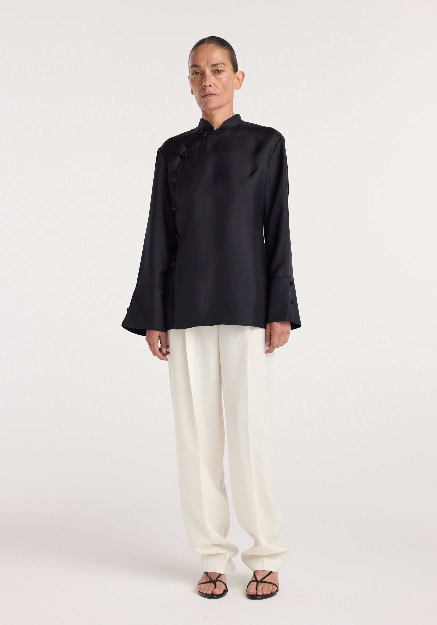 A woman wearing a black Pankou closure silk top | black with a high collar and asymmetric button detail paired with loose white trousers and black open-toe sandals.