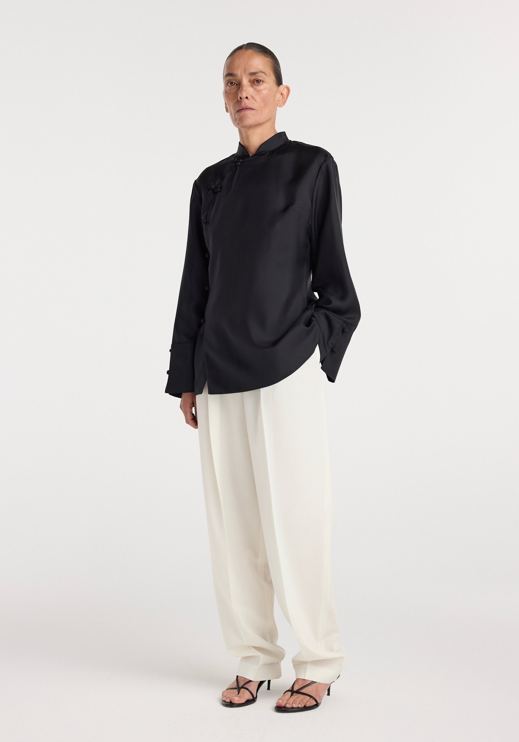 A model wearing a black Pankou closure silk top | black with long sleeves and mandarin collar, paired with loose white pants and black strappy heels against a plain white background.