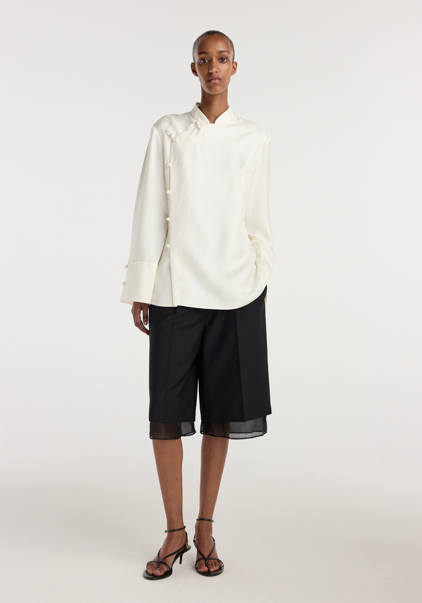 A model wearing a cream-colored Pankou closure silk top with long sleeves, paired with black knee-length shorts and black strappy heels, standing against a plain white background.