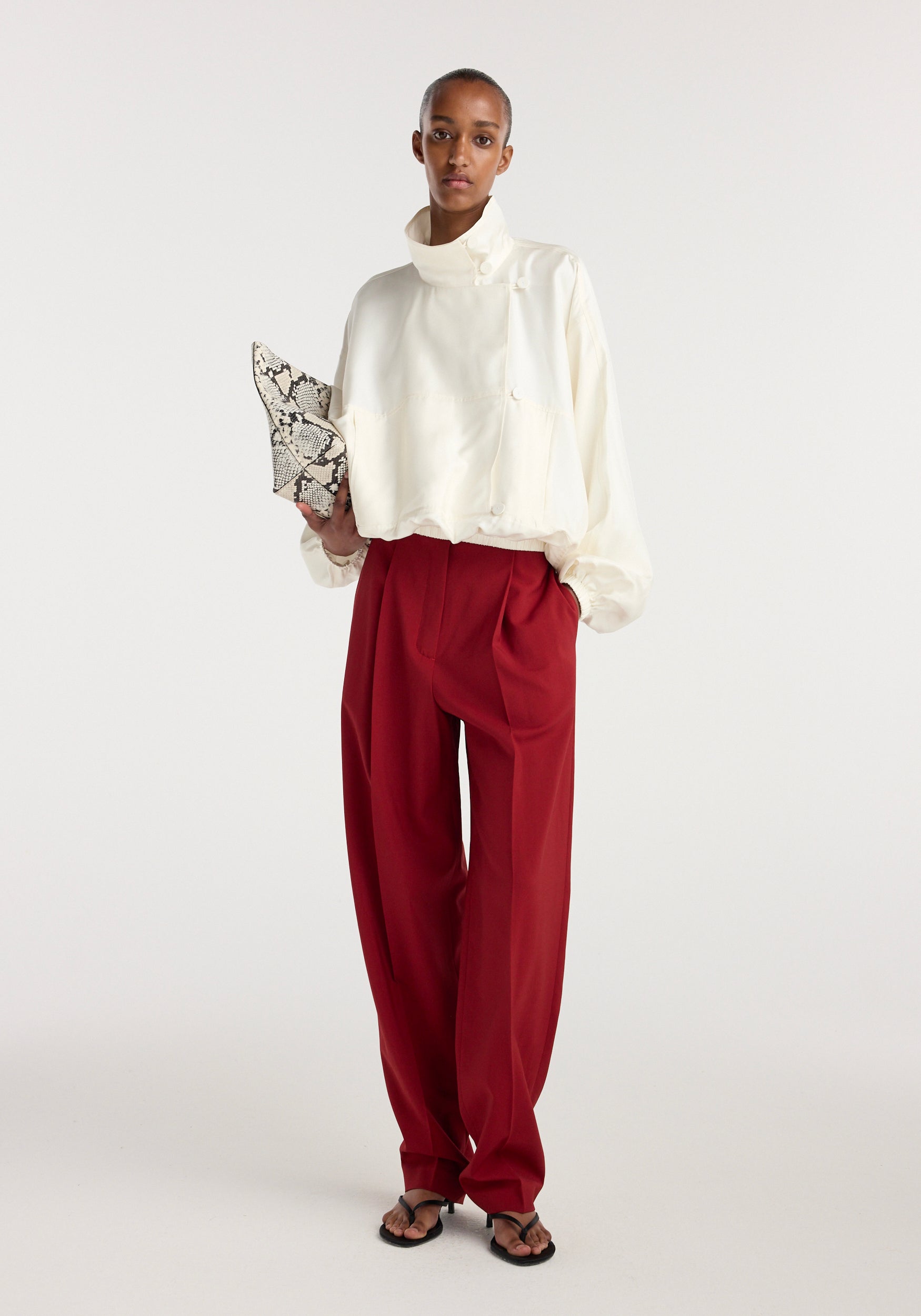 Model wearing a cream silk blouson anorak with a high collar and loose sleeves, paired with wide-legged red trousers, holding a patterned clutch against a plain background.