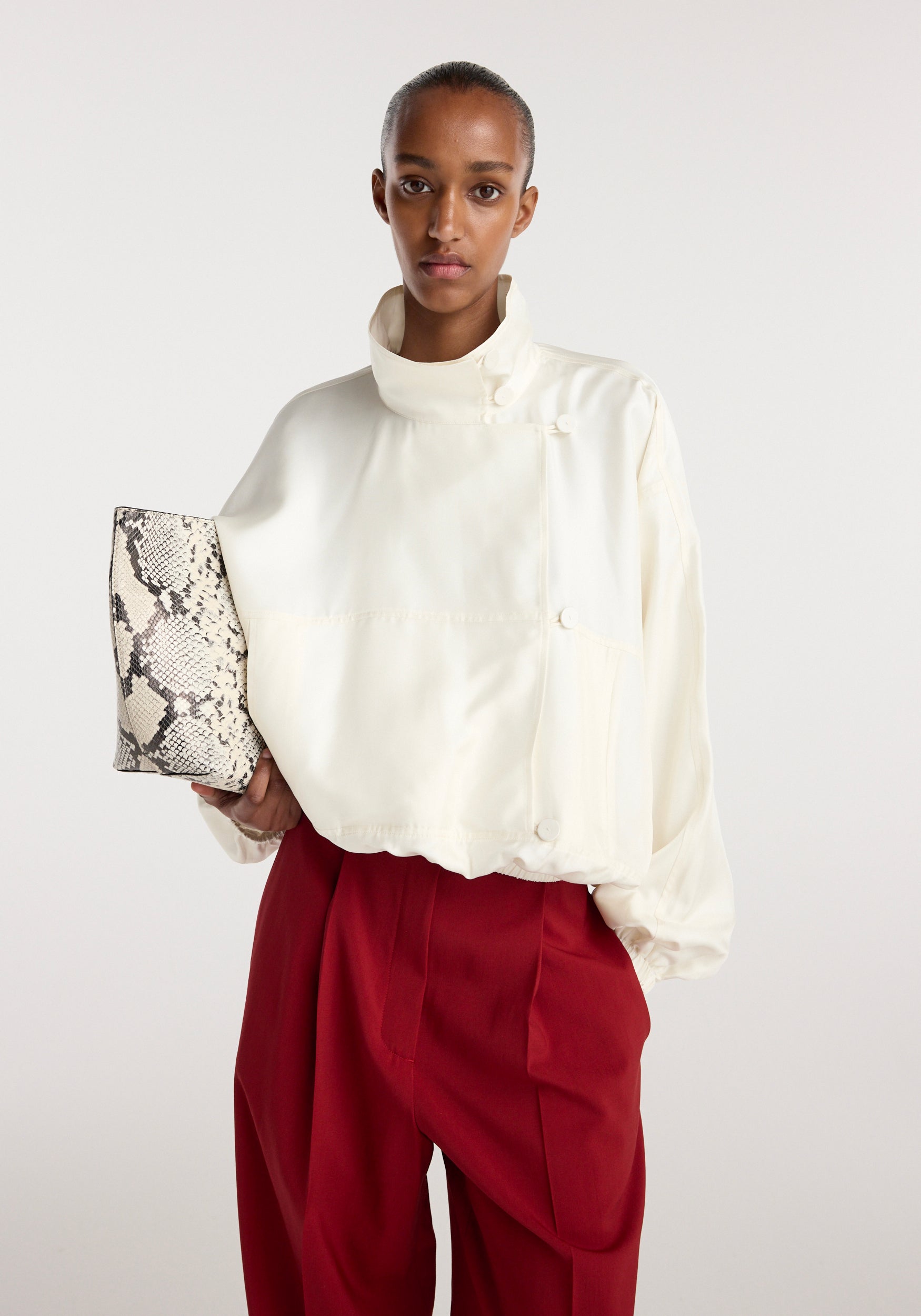 Model wearing a cream Silk blouson anorak with a high collar and side buttons, paired with red trousers, holding a snakeskin-patterned clutch bag.