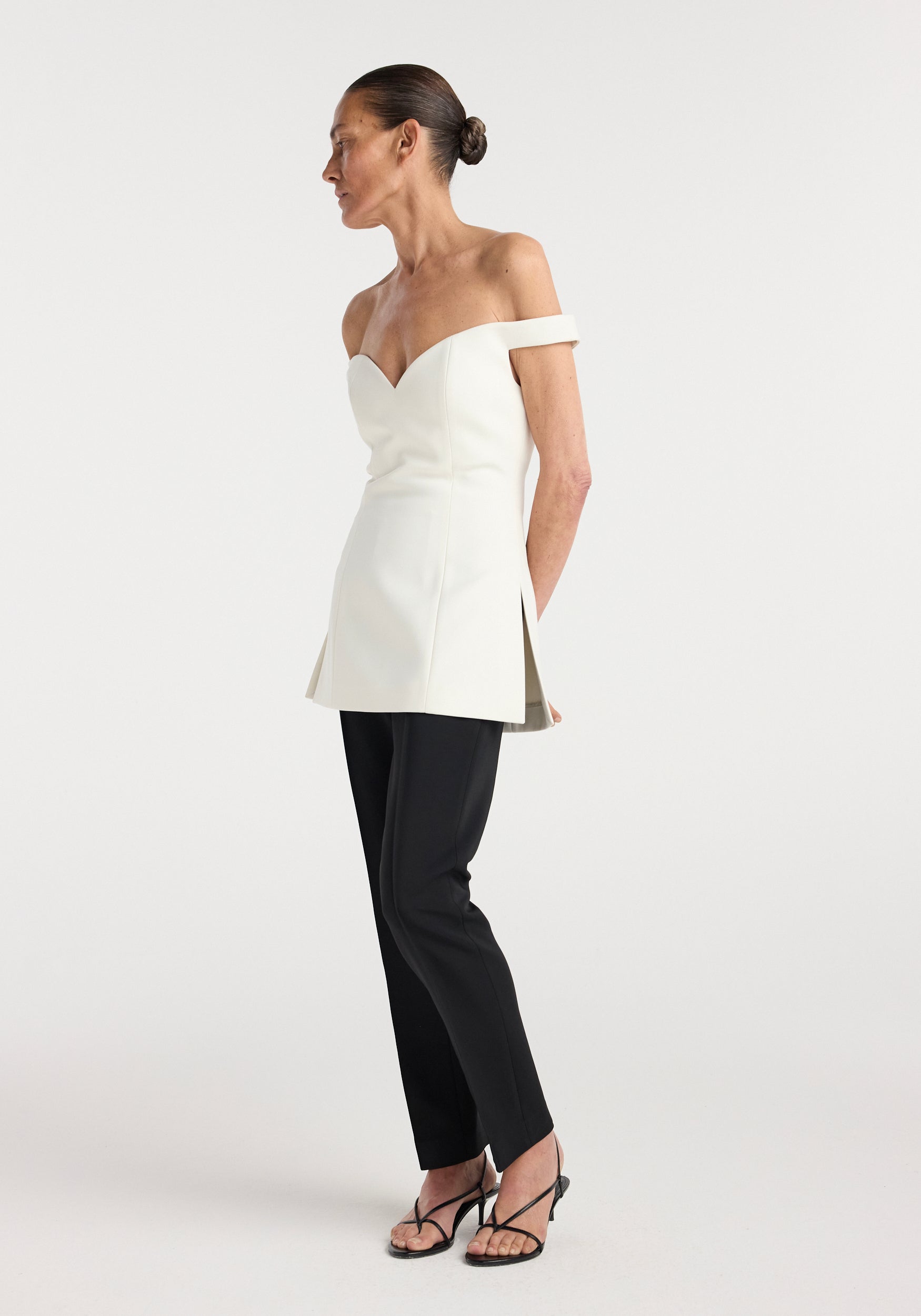 Woman wearing an off shoulder top | cream with a fitted bodice and slight flare at the hips, paired with black slim pants and black heeled sandals, standing in profile.