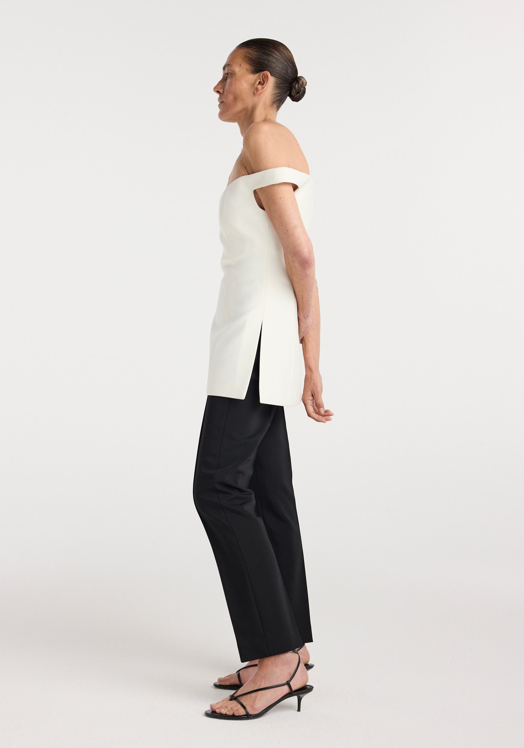 Woman wearing a cream off shoulder top with side slits paired with black straight-leg pants and black strappy heels, standing in profile against a plain background.