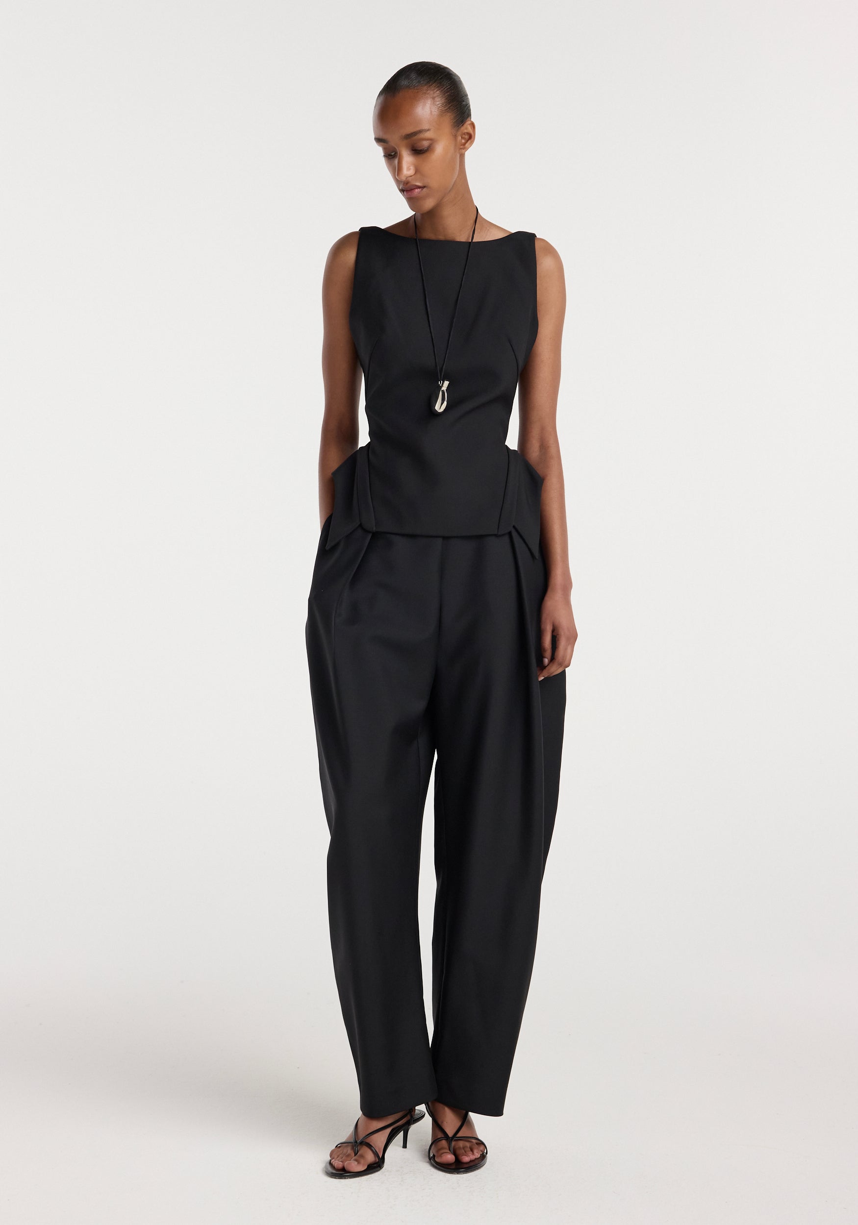 Model wearing a sleeveless black tailored top with a unique collar design paired with high-waisted loose black trousers and black strappy heels in a studio setting, featuring the Deconstructed collar top | black.