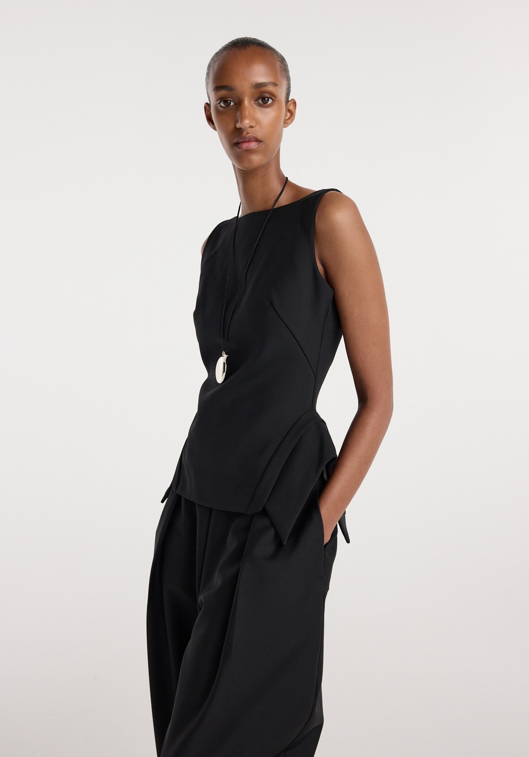 Model wearing a sleeveless black top with a unique deconstructed collar design and asymmetrical hem, styled with loose black pants and a long pendant necklace, "Deconstructed collar top | black".