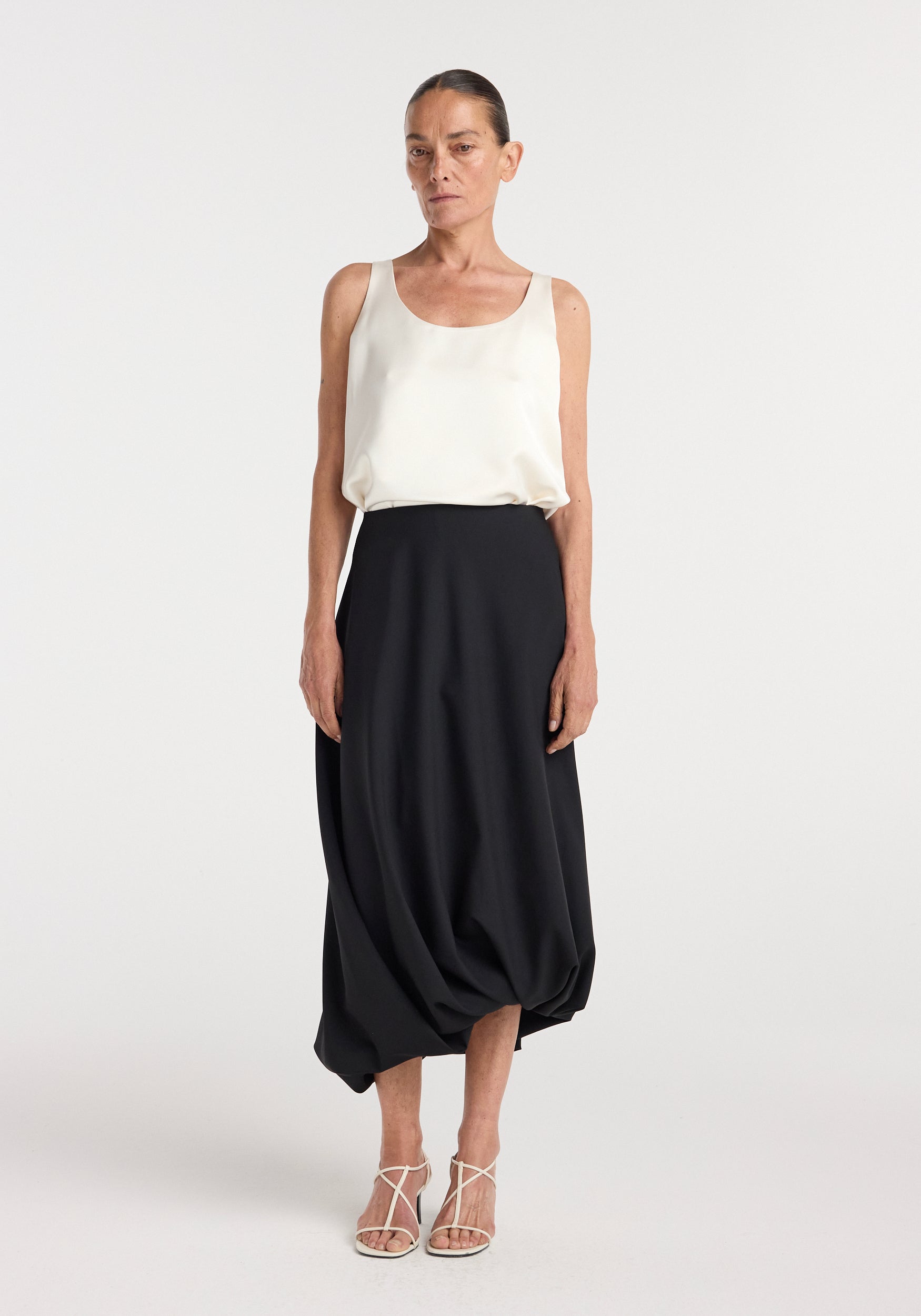 Woman wearing a white sleeveless top paired with a black asymmetrical twisted skirt | black and white strappy sandals against a plain white background.