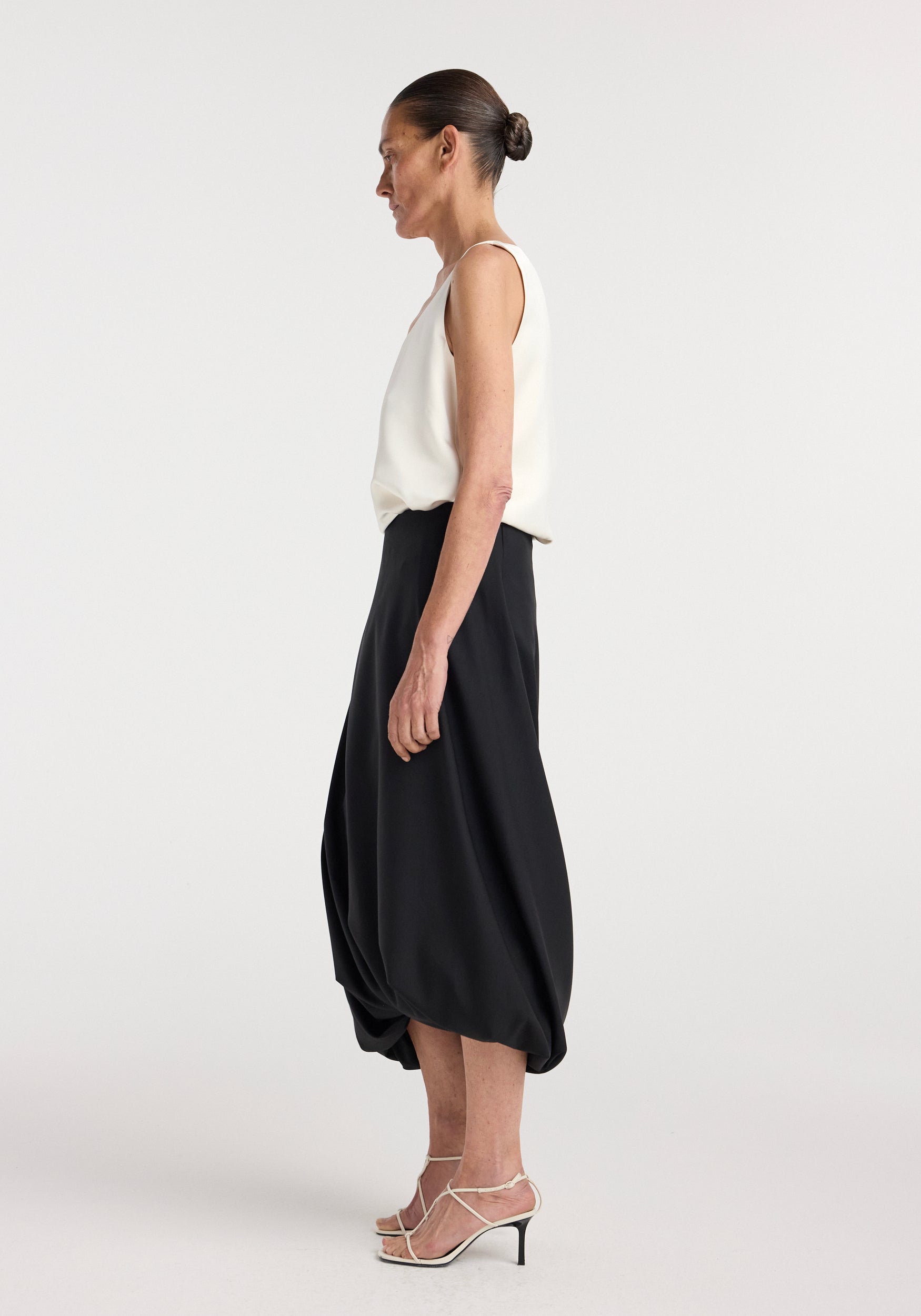 Side profile of a woman wearing a sleeveless white top paired with a black Twisted skirt | black featuring a draped, twisted hem design, styled with white strappy heels.