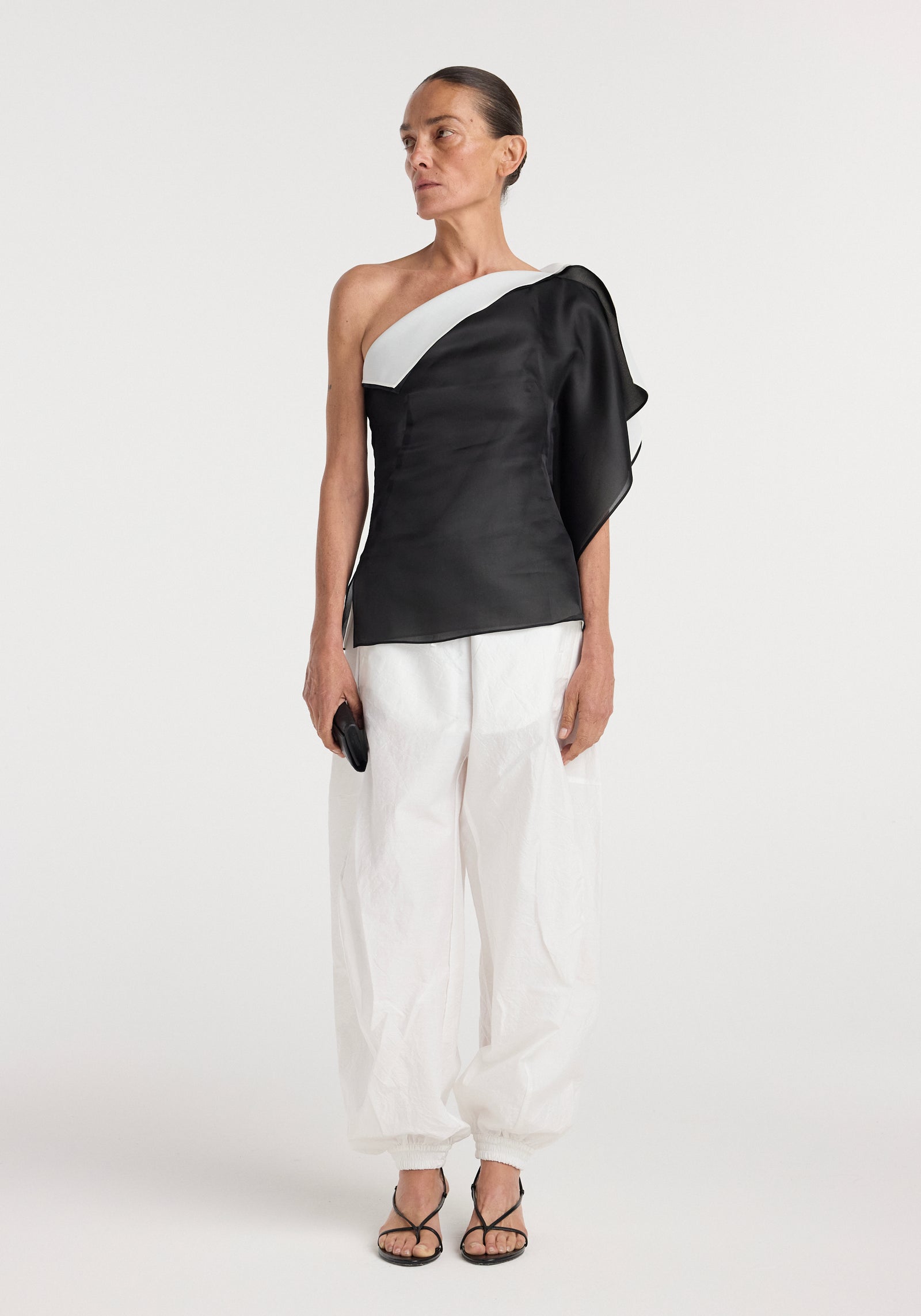 Woman wearing a sculptural double layered top | black with an asymmetrical neckline and layered design paired with loose white pants and black sandals, holding a small clutch.