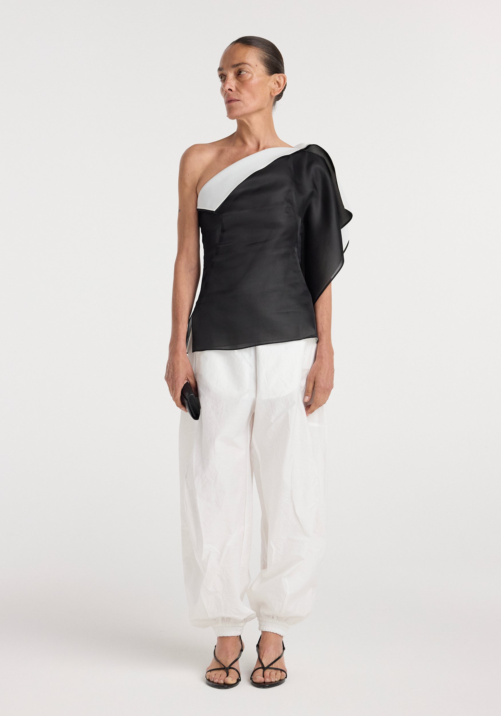 Woman wearing a sculptural double layered top | black with an asymmetrical neckline and layered design paired with loose white pants and black sandals, holding a small clutch.