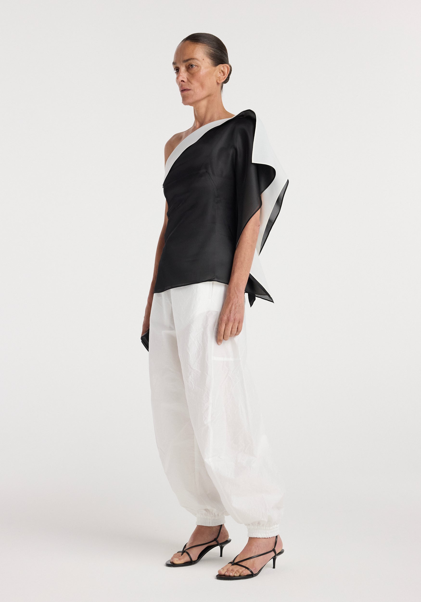 Woman wearing a sculptural double layered top | black with an asymmetrical design paired with loose white pants and black strappy heels, standing against a plain background.