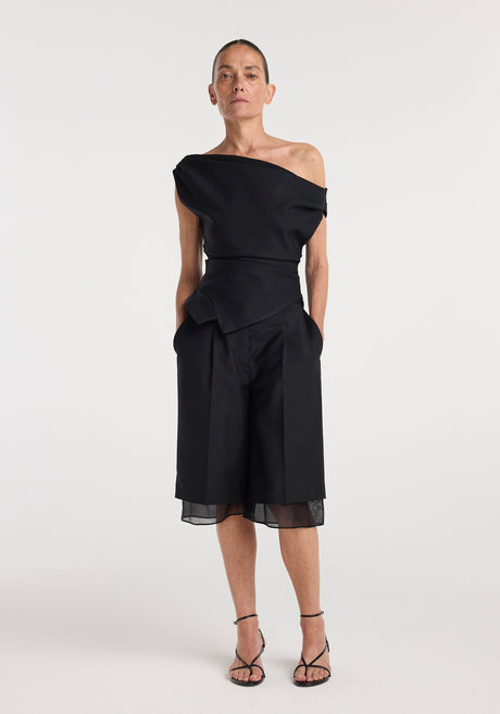Woman wearing a black off shoulder lapel top with an asymmetrical neckline paired with black knee-length wide-leg shorts and strappy black heels, standing against a plain white background.