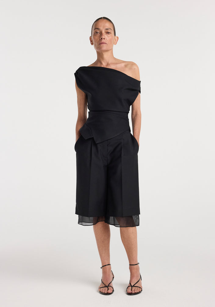 Woman wearing a black off shoulder lapel top with an asymmetrical neckline paired with black knee-length wide-leg shorts and strappy black heels, standing against a plain white background.