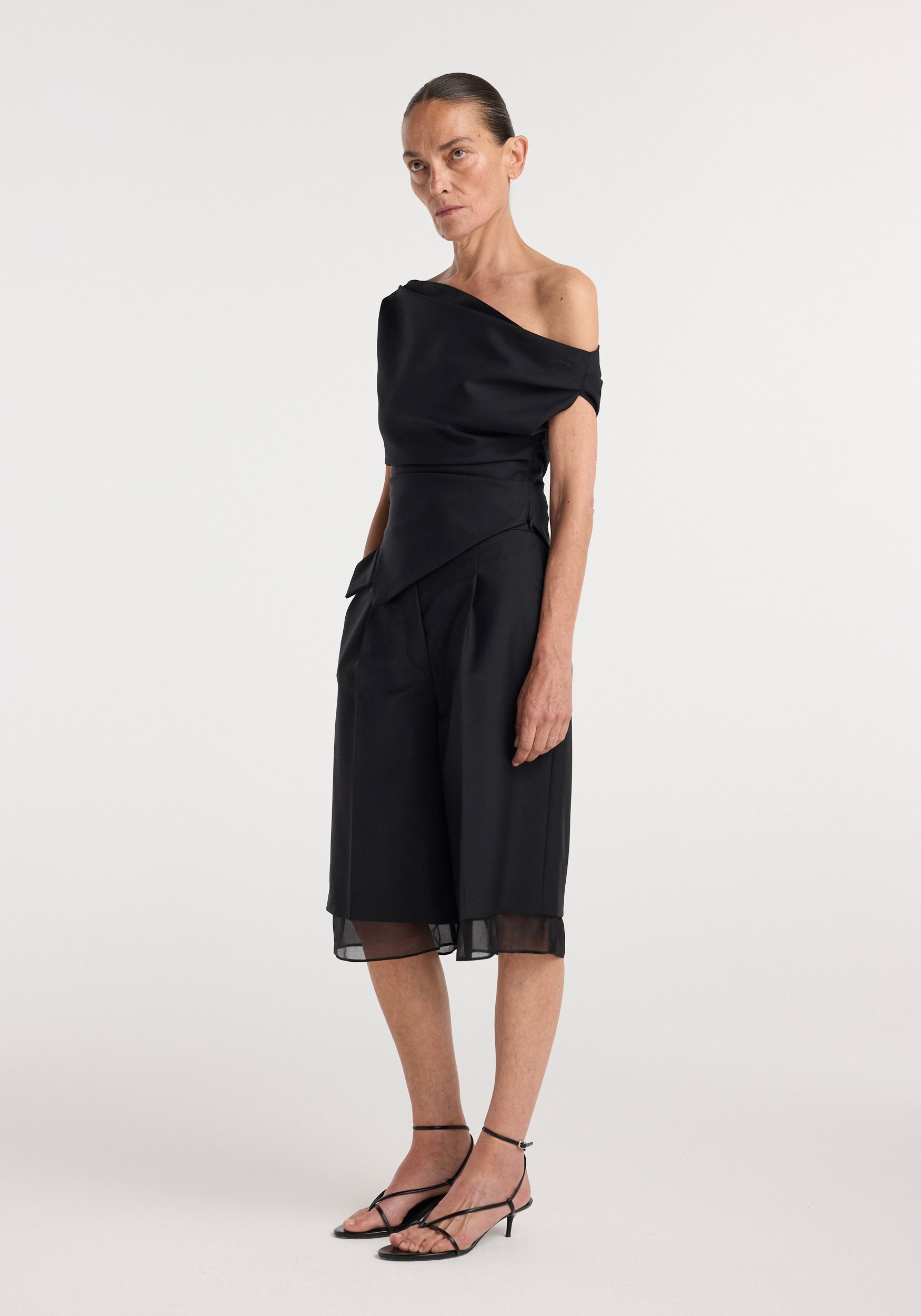 Woman wearing a black off shoulder lapel top | black paired with wide-leg culottes and strappy black heels, standing against a plain white background.