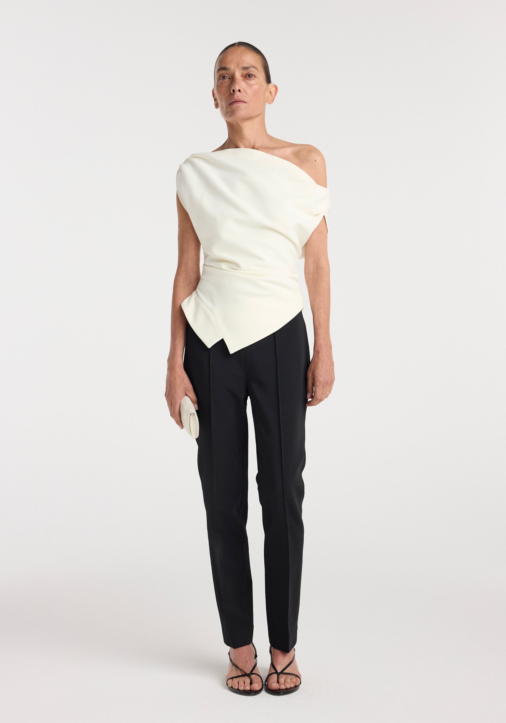 Woman wearing an off shoulder lapel top | off white paired with black tailored pants and black strappy heels, holding a small light-colored clutch against a plain white background.