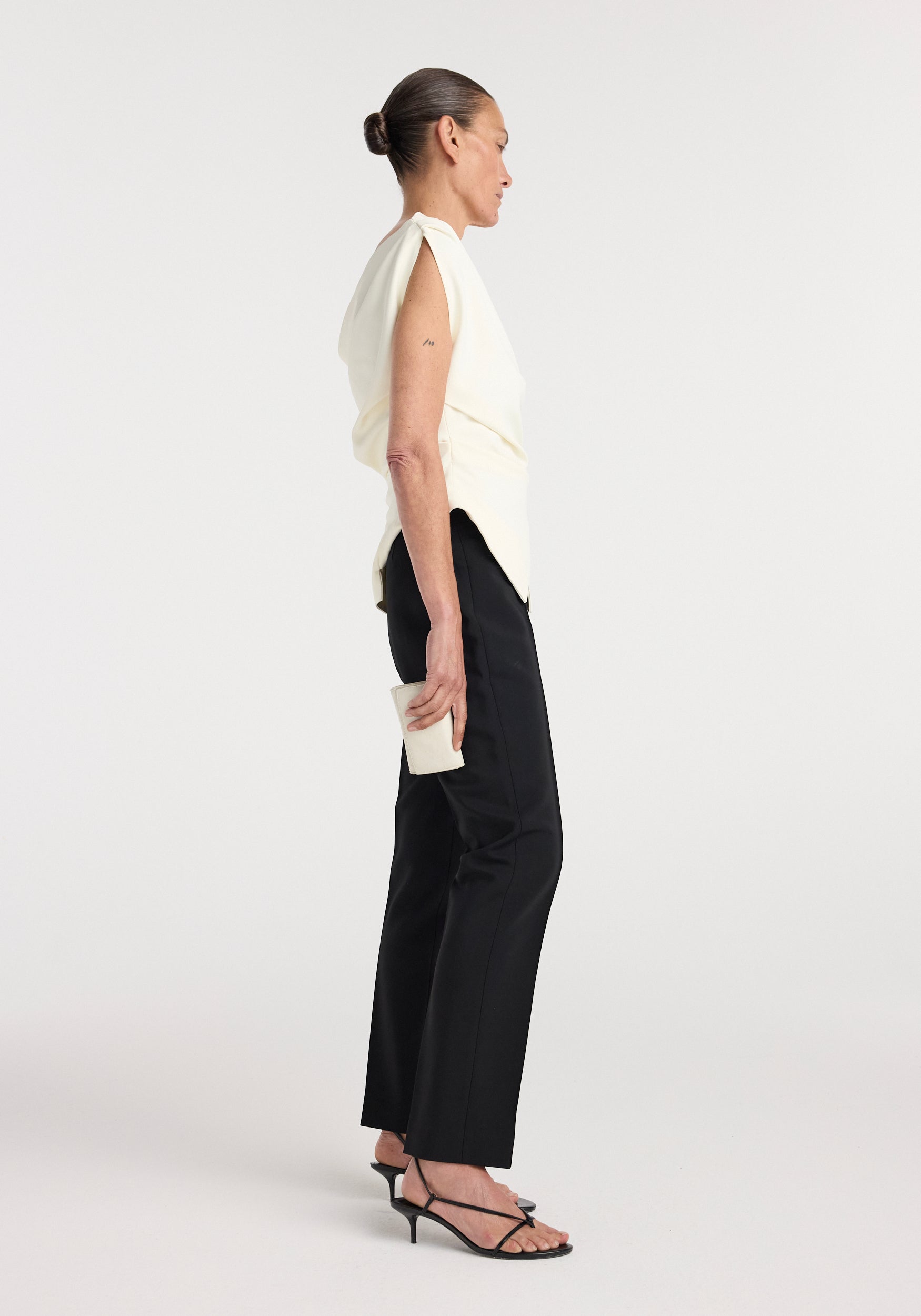 Side view of a woman wearing an off shoulder lapel top | off white with black tailored pants and black strappy heeled sandals, holding a small white clutch.