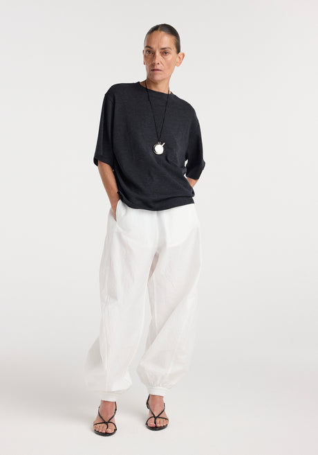 Woman wearing an oversized merino t-shirt | anthracite melange with half sleeves, paired with loose white pants and black strappy sandals, standing against a plain background.