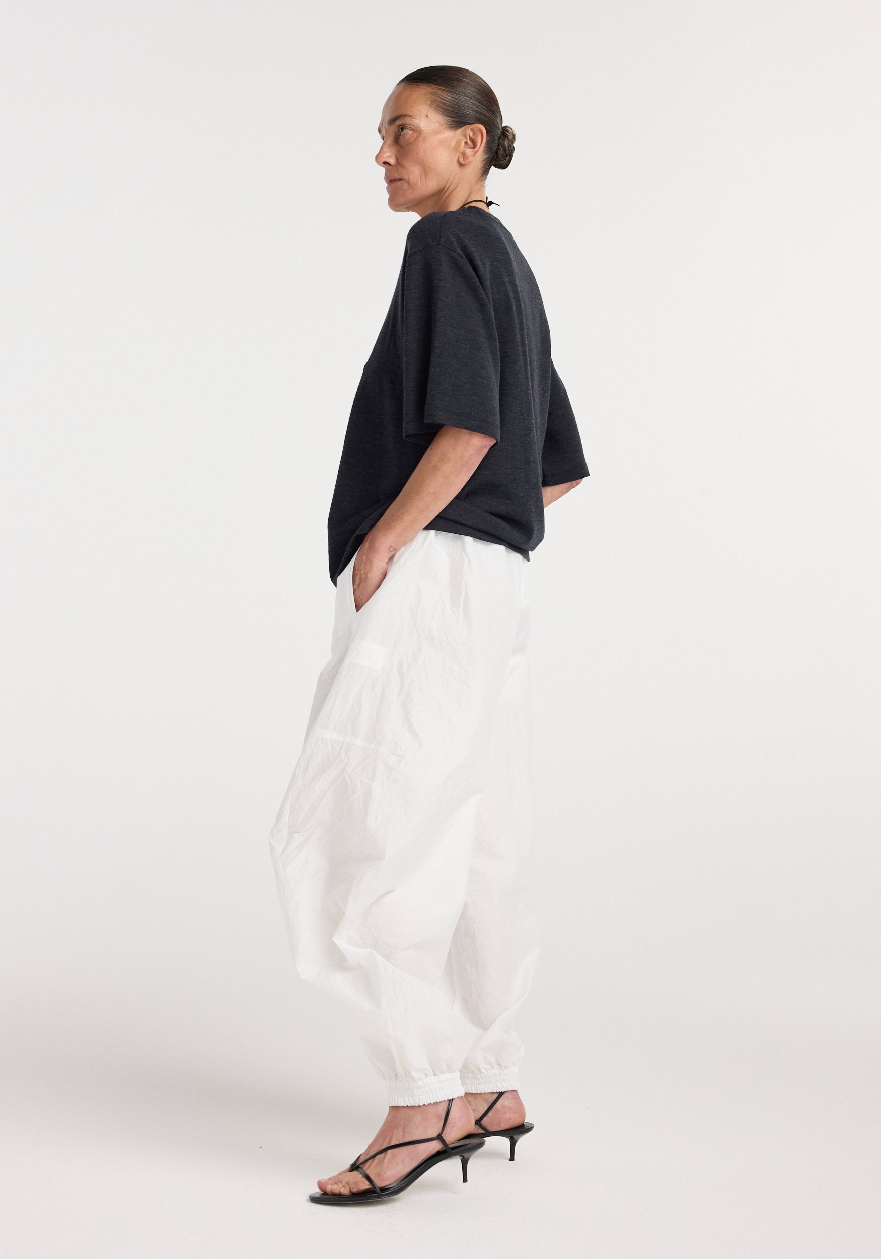 Woman wearing an oversized merino t-shirt | anthracite melange paired with loose white pants and black strappy heels, standing in profile against a plain background.