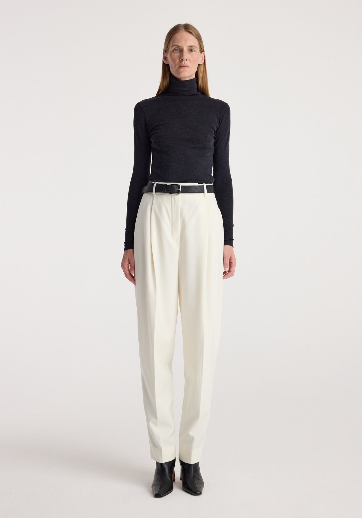 Woman wearing a fine merino turtleneck | anthracite melange paired with high-waisted white pants, black belt, and black ankle boots against a plain background.