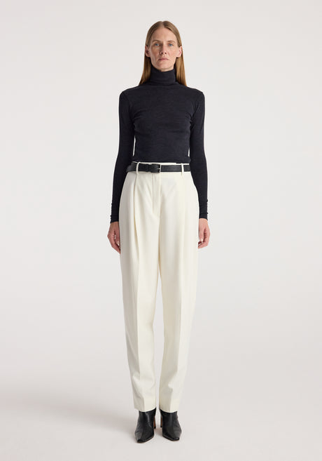 Woman wearing a fine merino turtleneck | anthracite melange paired with high-waisted white pants, black belt, and black ankle boots against a plain background.