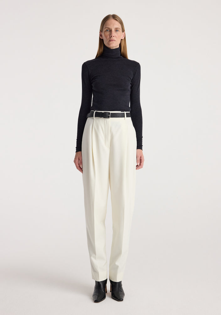 Woman wearing a fine merino turtleneck | anthracite melange paired with high-waisted white pants, black belt, and black ankle boots against a plain background.