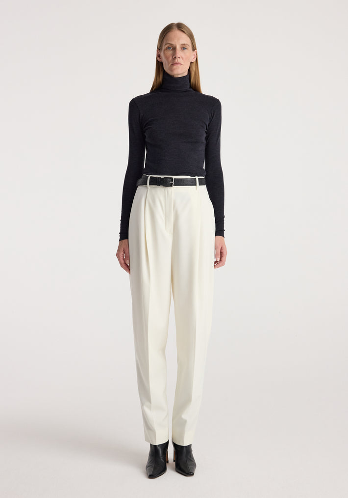 Woman wearing a fine merino turtleneck | anthracite melange paired with high-waisted white pants, black belt, and black ankle boots against a plain background.