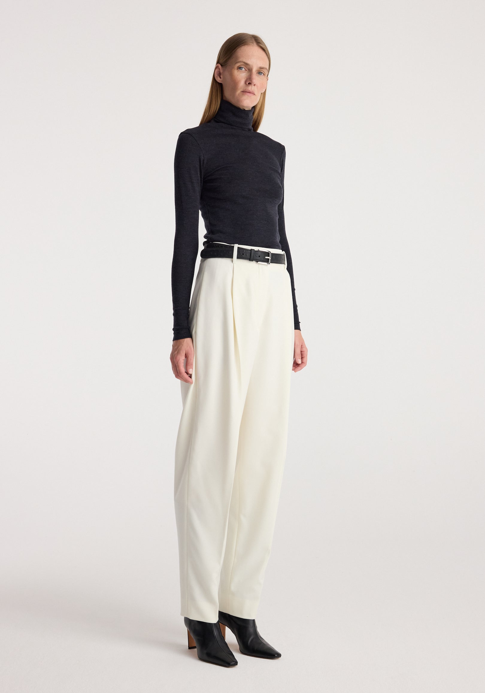 Woman wearing a fitted fine merino turtleneck | anthracite melange paired with high-waisted white trousers and black ankle boots against a plain background.