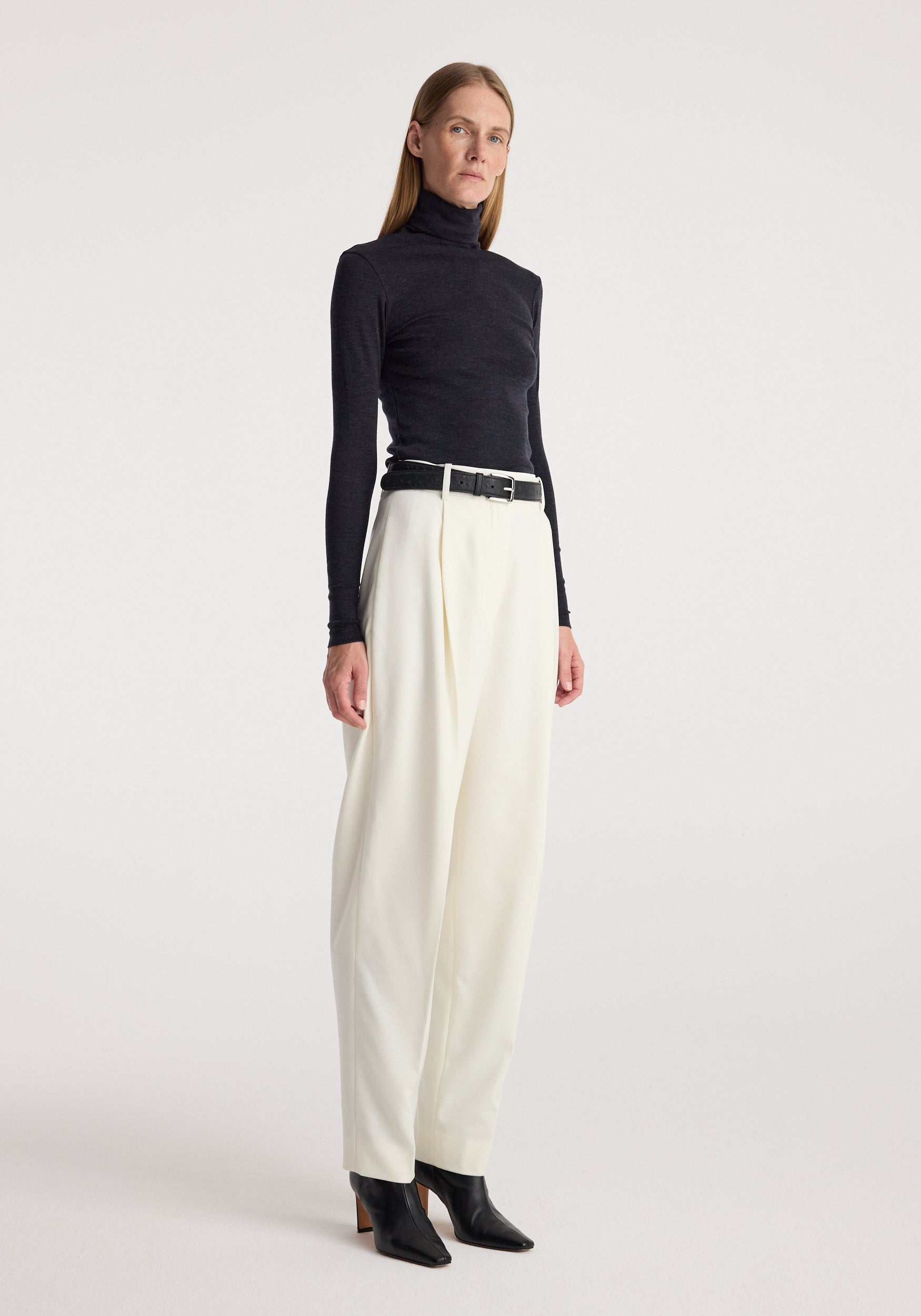Woman wearing a fitted fine merino turtleneck | anthracite melange paired with high-waisted white trousers and black ankle boots against a plain background.