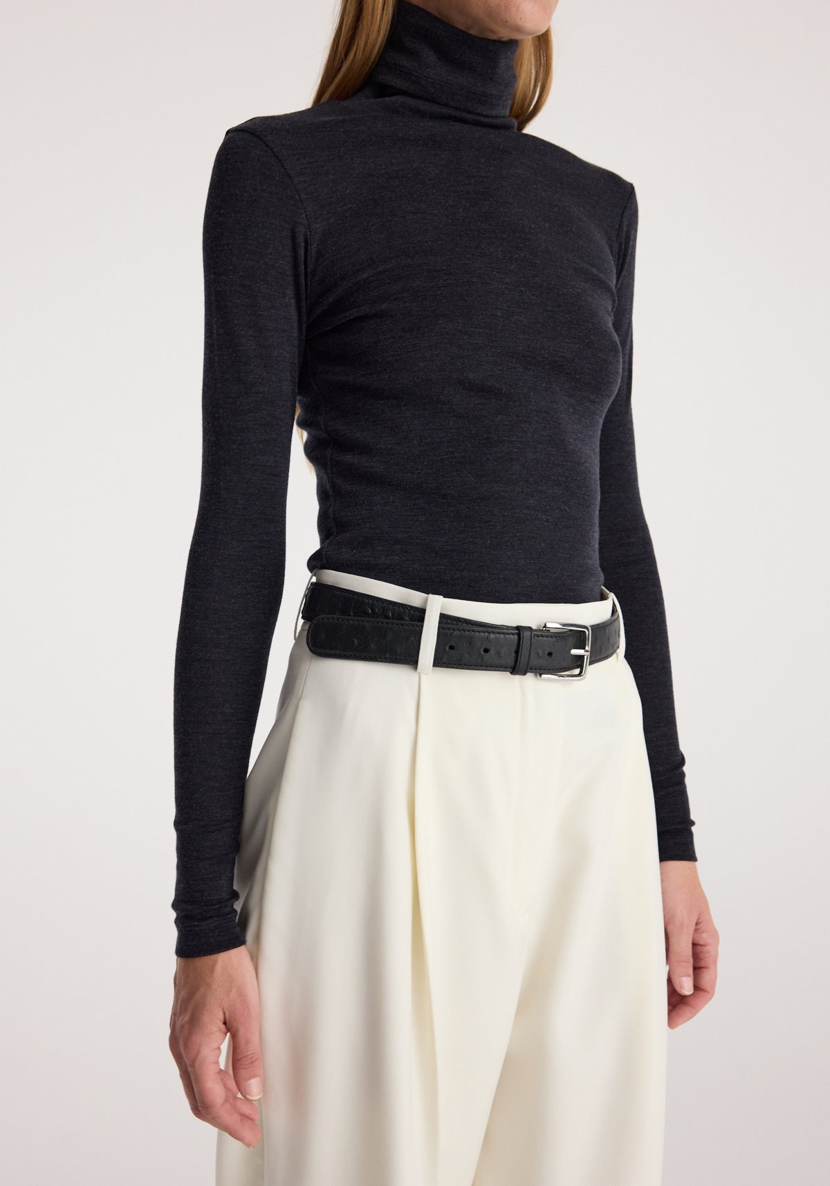 Woman wearing a fitted Fine merino turtleneck | anthracite melange in dark grey paired with high-waisted cream trousers and a black belt, shown from neck to hips.