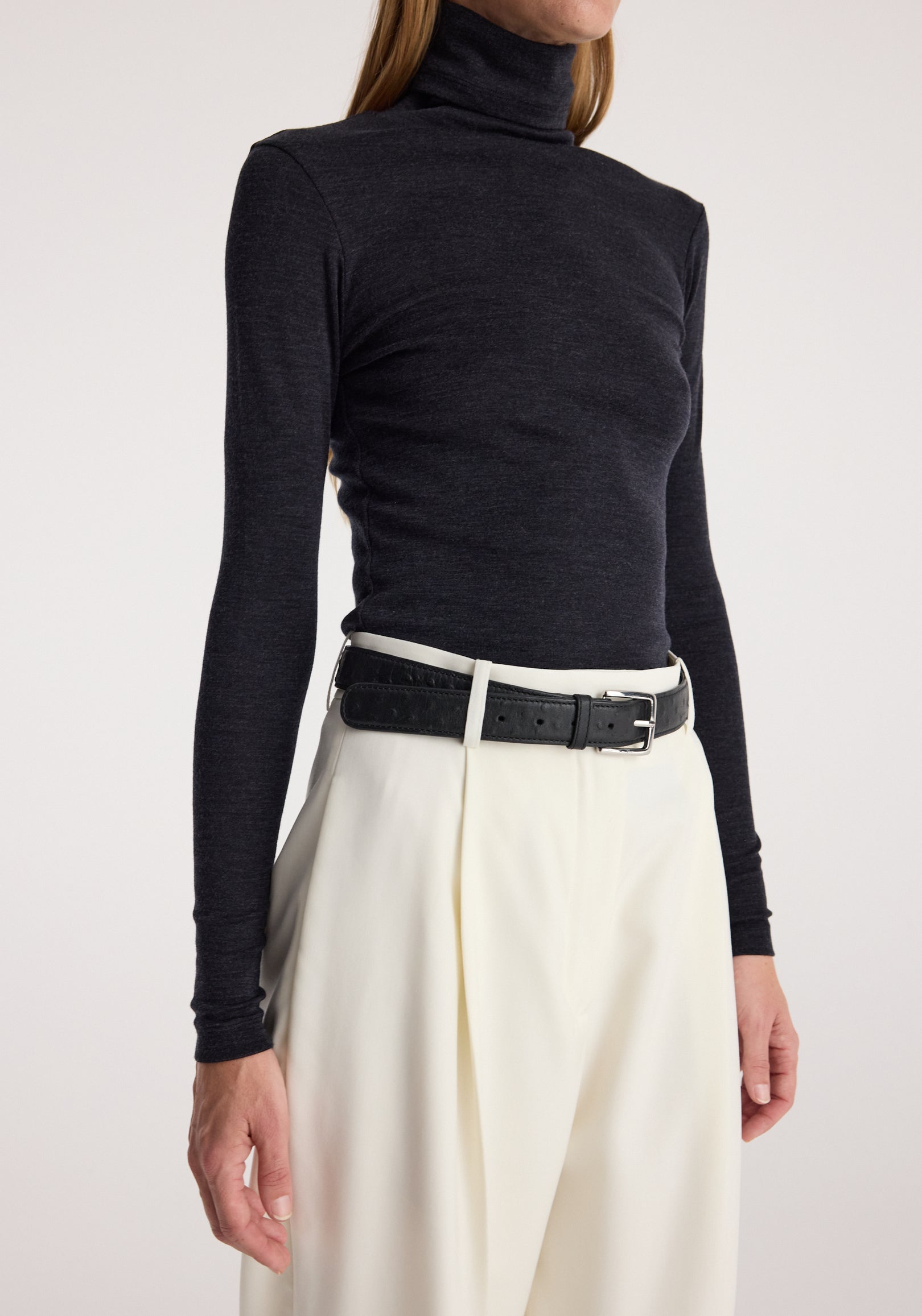 Woman wearing a fitted Fine merino turtleneck | anthracite melange in dark grey paired with high-waisted cream trousers and a black belt, shown from neck to hips.