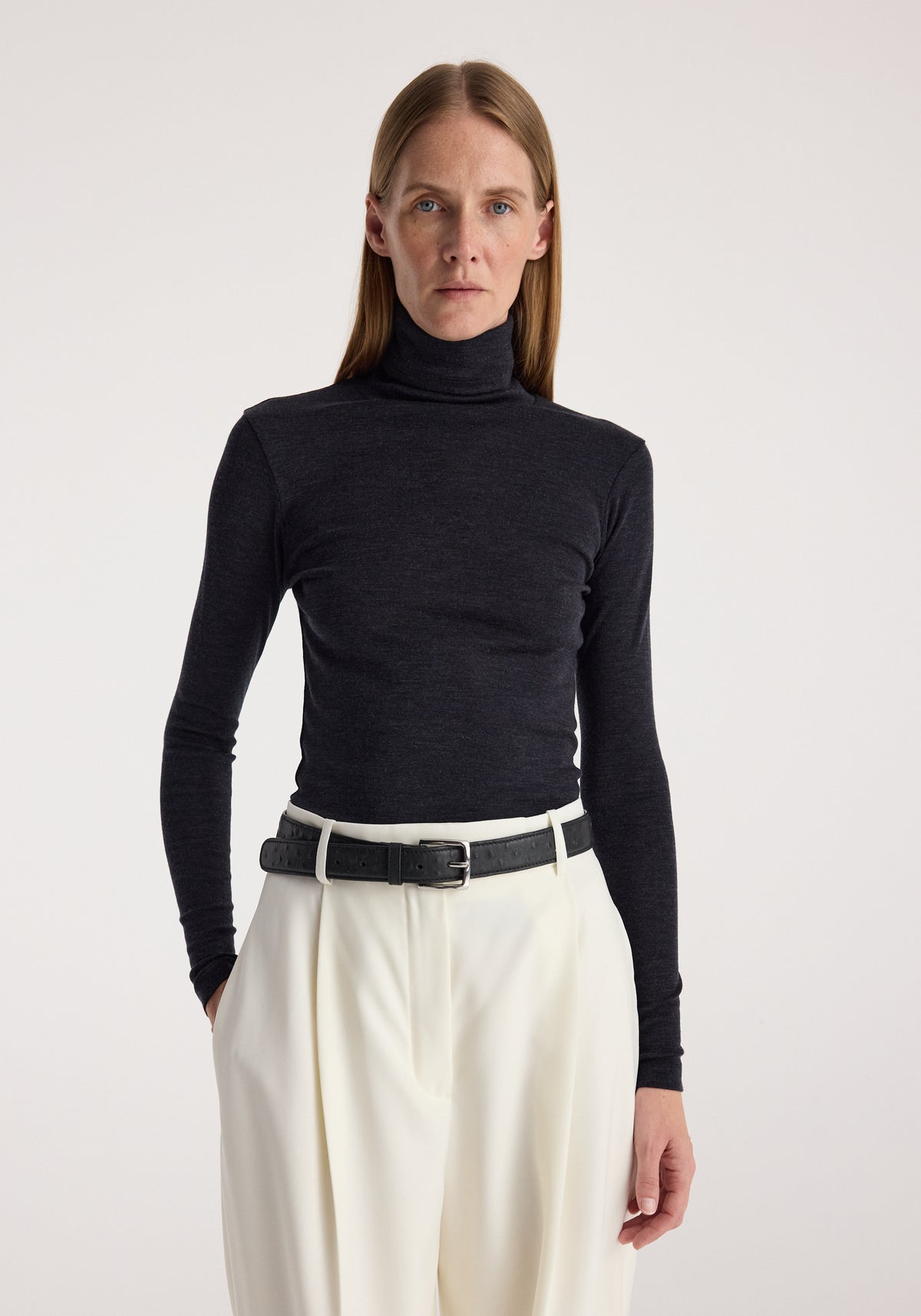 Woman wearing a fitted fine merino turtleneck | anthracite melange paired with high-waisted white trousers and a black belt, standing against a plain background.