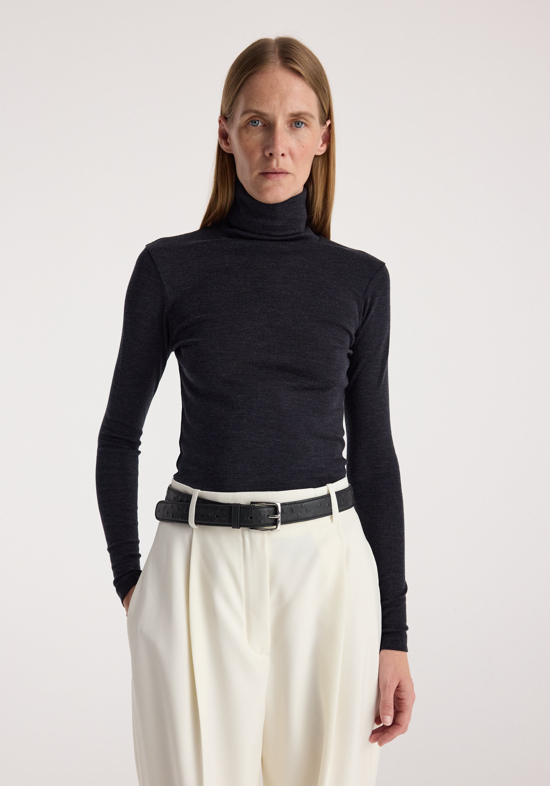 Woman wearing a fitted fine merino turtleneck | anthracite melange paired with high-waisted white trousers and a black belt, standing against a plain background.