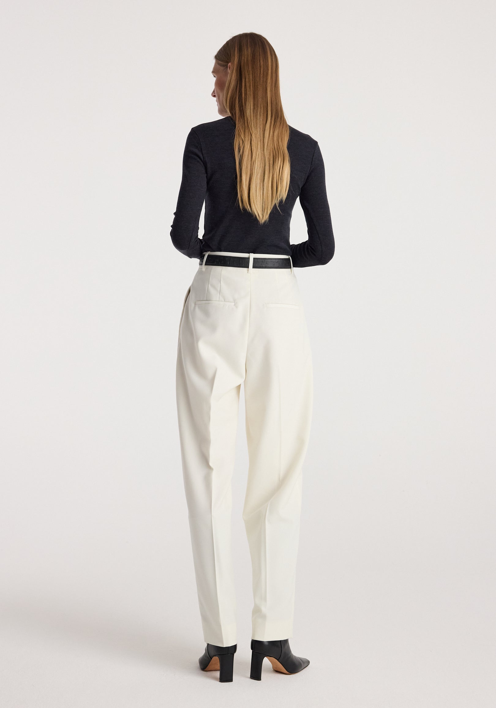 Woman seen from behind wearing a Fine merino turtleneck | anthracite melange paired with white trousers and black heeled ankle boots, standing against a plain background.