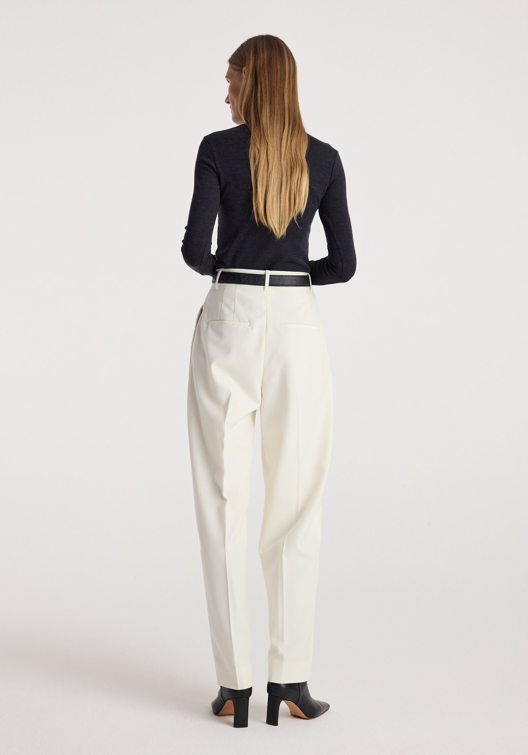 Woman seen from behind wearing a Fine merino turtleneck | anthracite melange paired with white trousers and black heeled ankle boots, standing against a plain background.