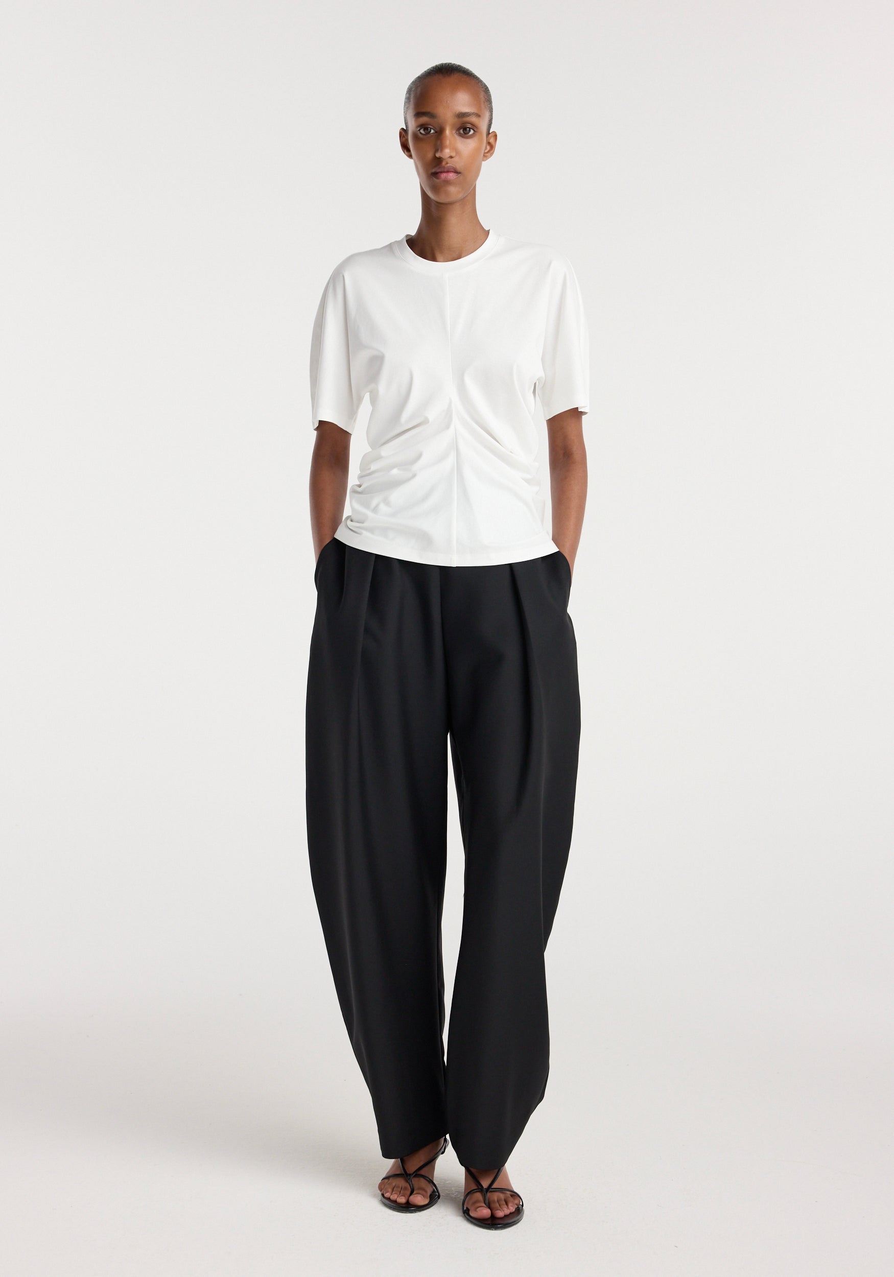 Model wearing a Draped t-shirt | off white paired with loose black trousers and black open-toe sandals, standing against a plain white background.